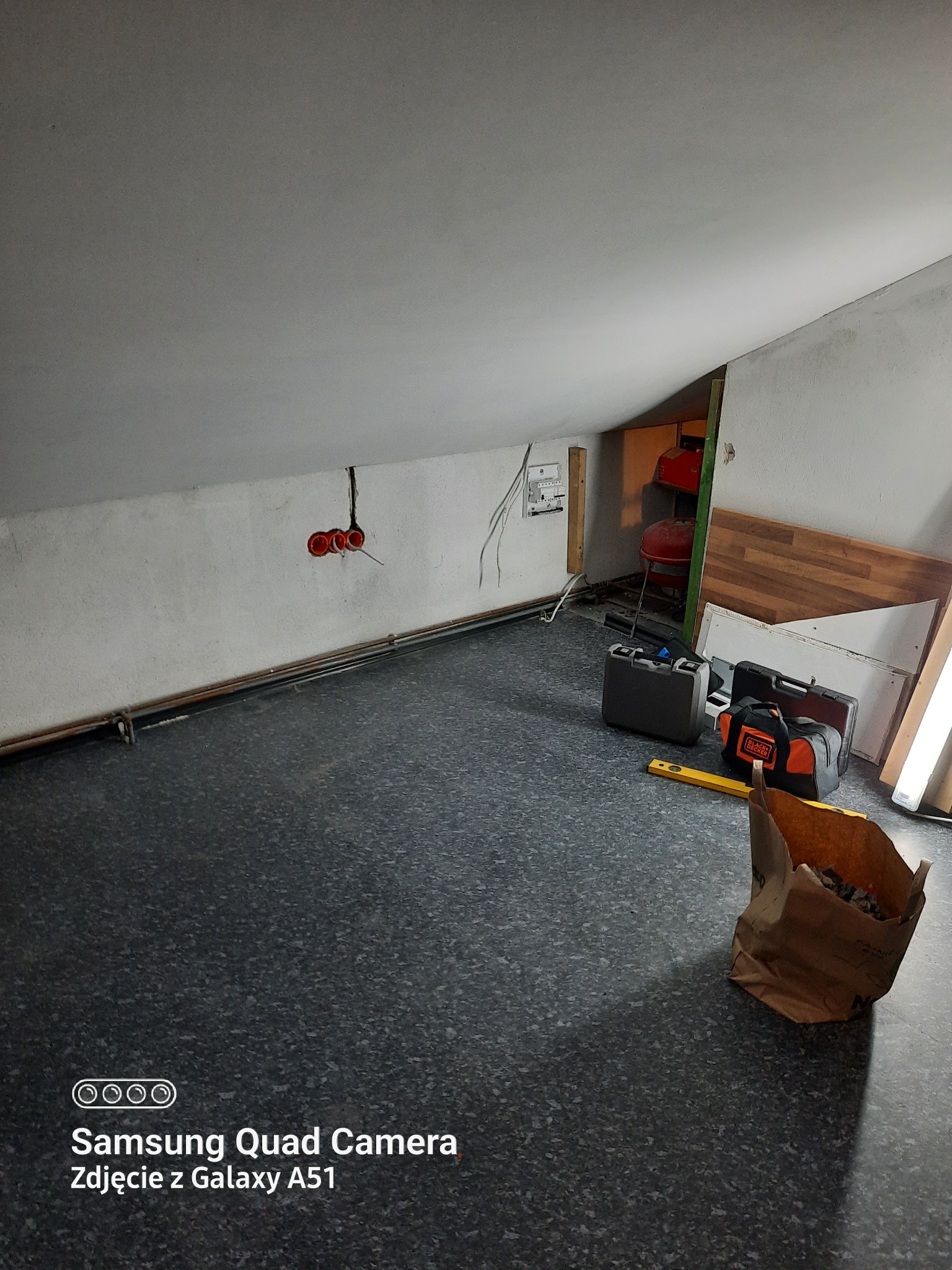 Poddasze w trakcie remontu z widocznymi puszkami instalacyjnymi w ścianie, torbą z odpadami, skrzynkami narzędziowymi i poziomnicą na podłodze.