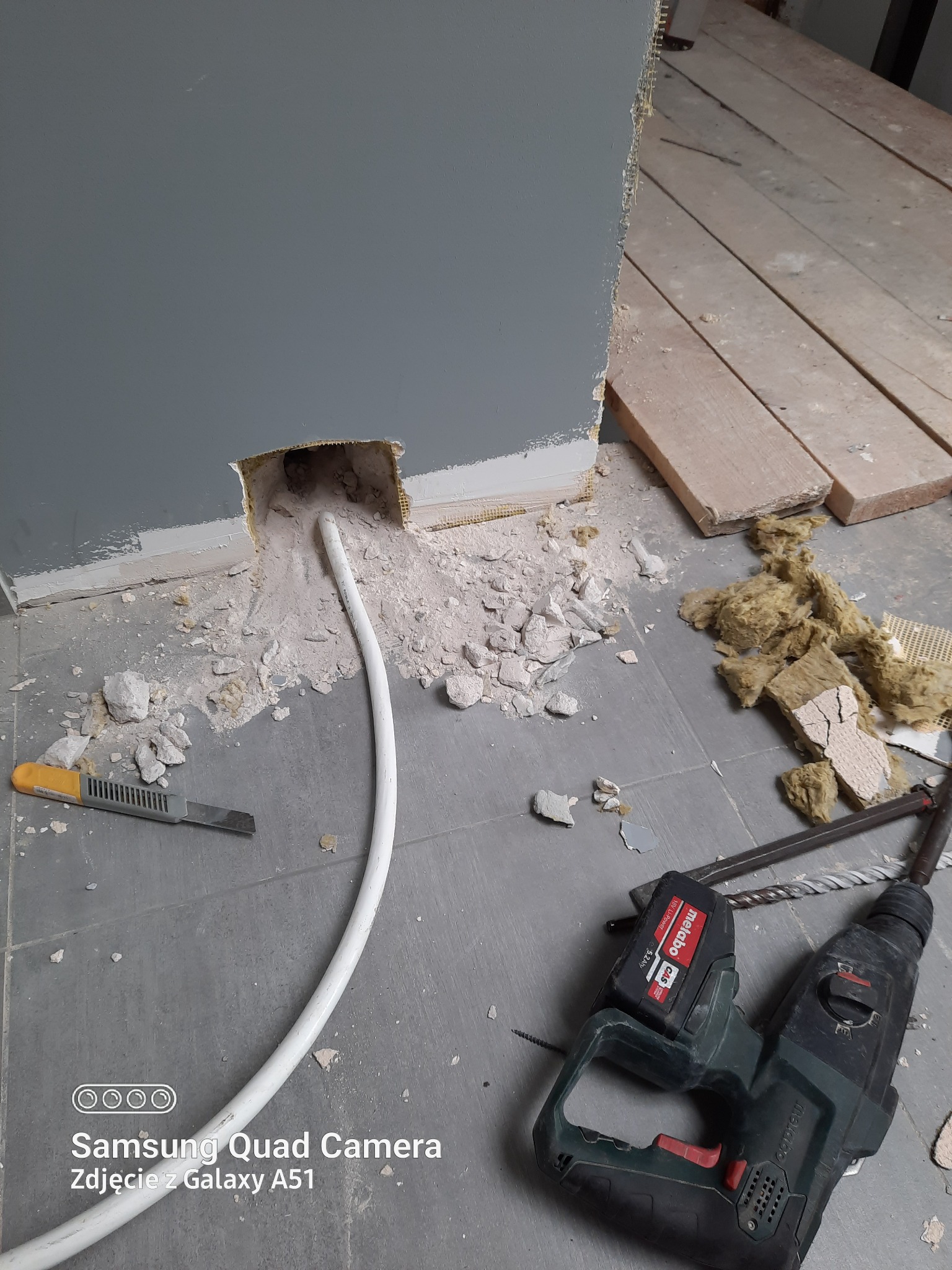 Otwór w ścianie z wystającym białym kablem, gruz i narzędzia do wiercenia na podłodze z płytek. Widoczny fragment izolacji i nóż do tapet.