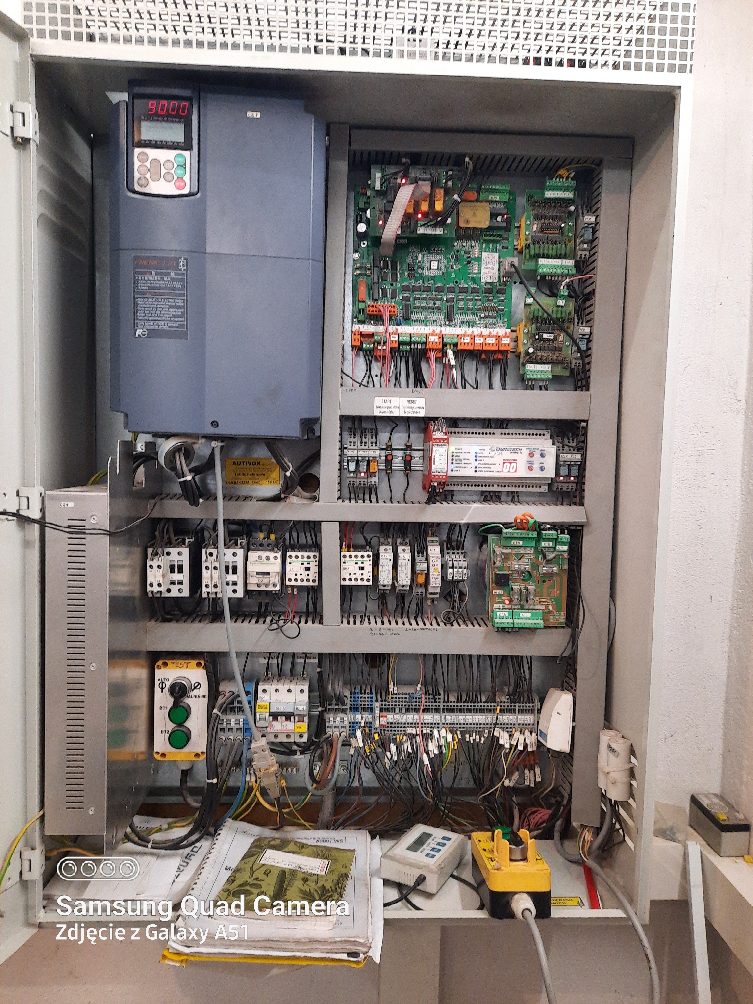Wnętrze szafy sterowniczej z widocznymi modułami elektronicznymi, przewodami i przyciskami sterującymi; na dole notatnik z zieloną okładką i miernik elektryczny.