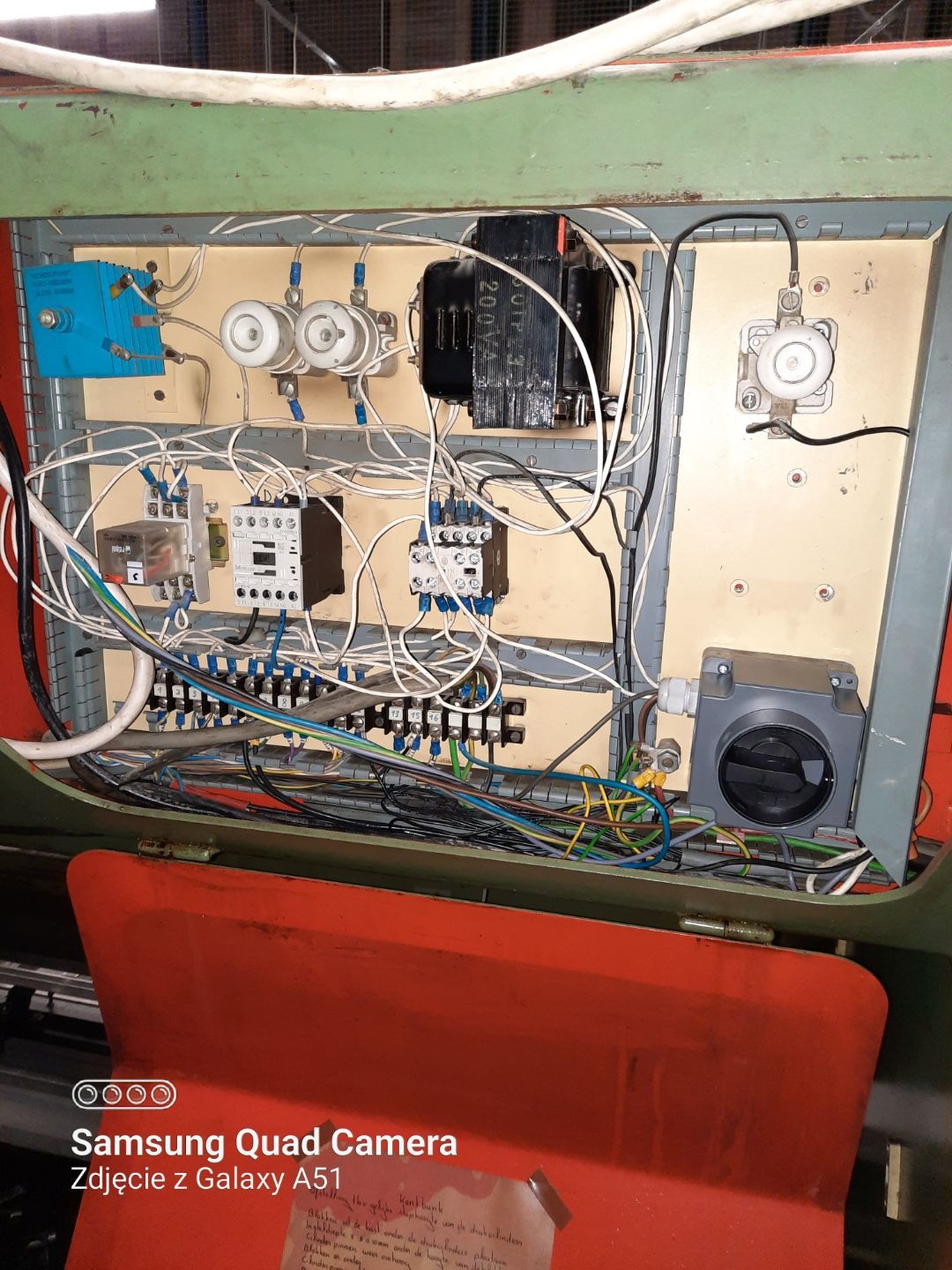 Otwarta skrzynka elektryczna z licznymi przewodami, transformatorem, bezpiecznikami i elementami sterującymi, widoczna kartka z odręcznym pismem w języku niderlandzkim przyklejona do obudowy.