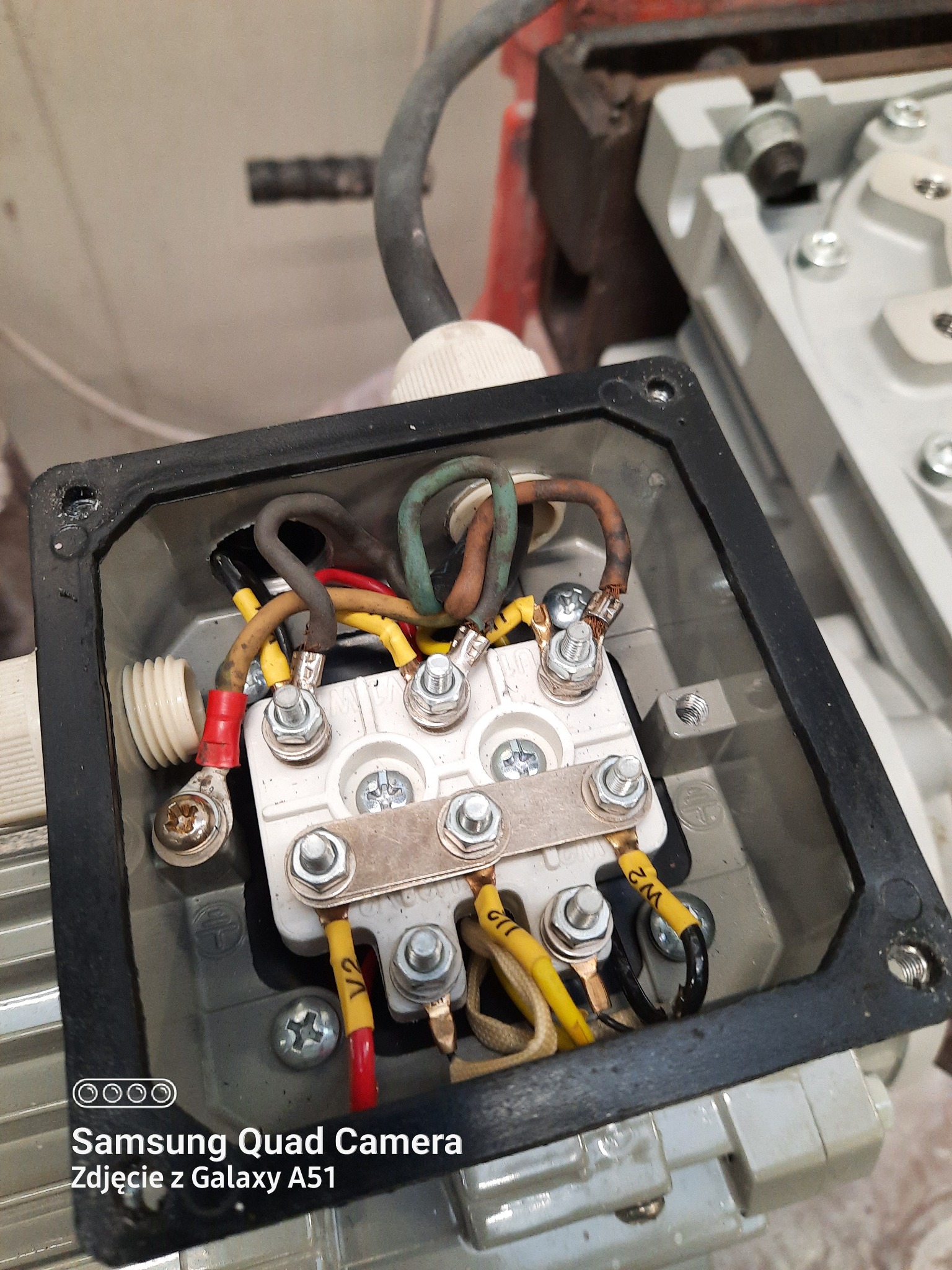 Przyłącze elektryczne silnika: widok z góry na listwę zaciskową z podłączonymi przewodami w kolorach żółtym, czerwonym, brązowym i zielonym, zabezpieczonymi nakrętkami. Czarna obudowa przyłącza...