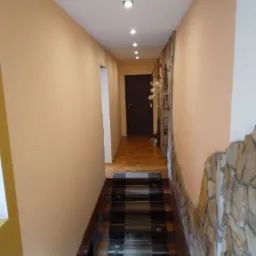 Korytarz z schodami z drewnianymi stopniami i szklanymi wstawkami, ściany w kolorze brzoskwini, fragment ściany wykończony kamieniem dekoracyjnym, drewniana podłoga w jodełkę, oświetlenie sufitowe...