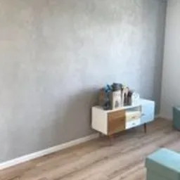 Ściana pokryta dekoracyjną, srebrzystą gładzią w odcieniach szarości, z widoczną strukturą i delikatnym połyskiem. Na pierwszym planie fragment podłogi z paneli w odcieniach jasnego drewna...