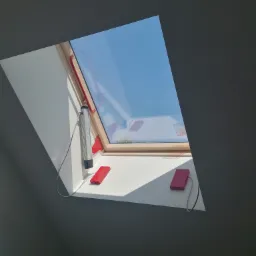 Zautomatyzowane okno dachowe z drewnianą ramą, widoczne elementy sterujące w kolorze czerwonym i szarym, błękitne niebo za oknem.