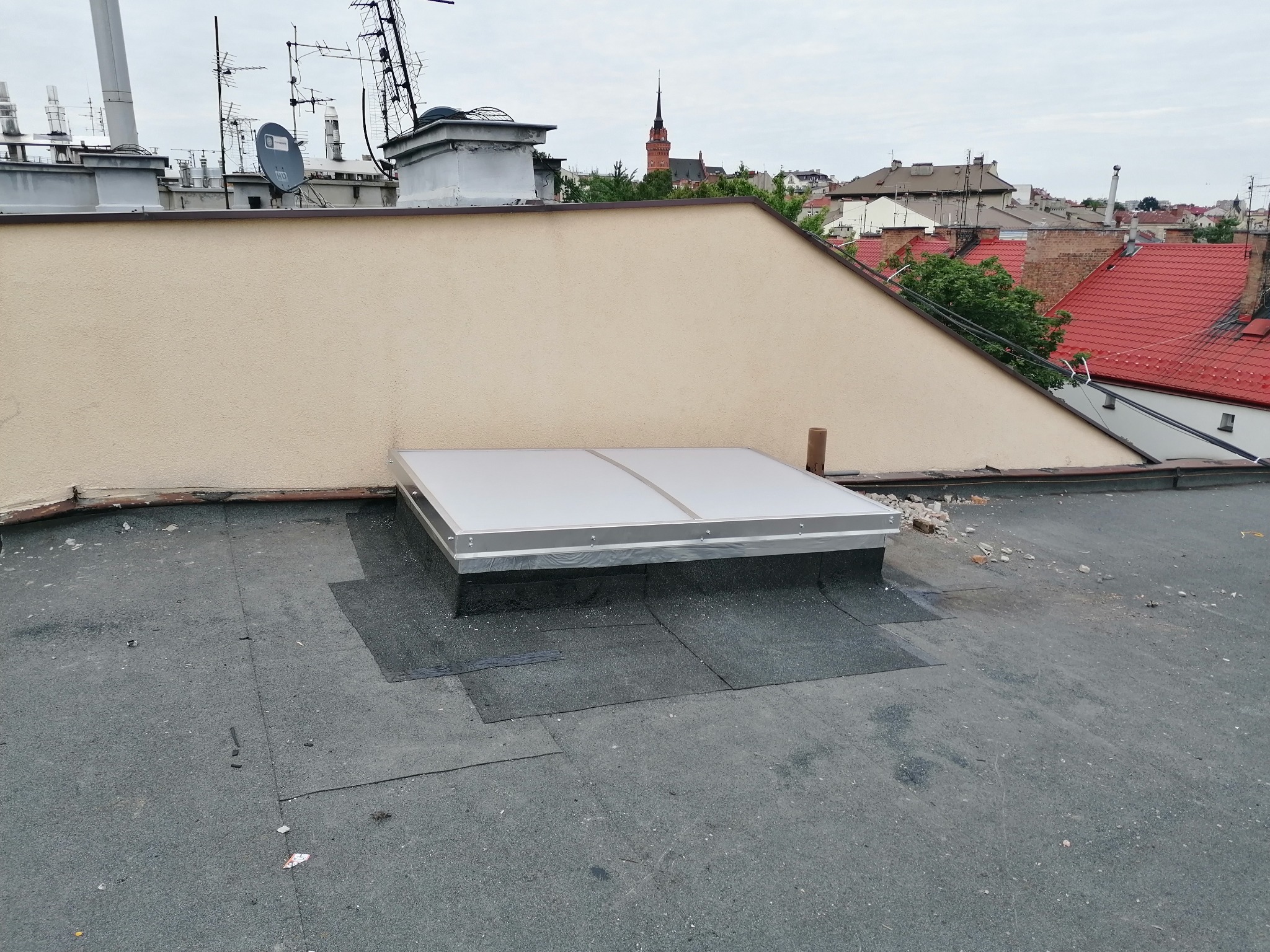 Płaski dach pokryty papą z zamontowanym świetlikiem dachowym; w tle zabudowa miejska z wieżą kościelną.