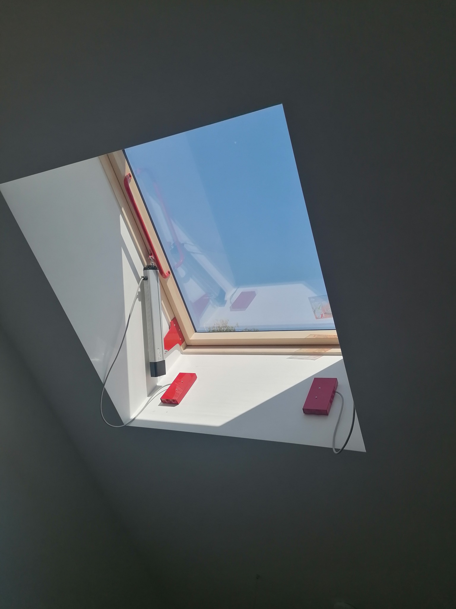 Zautomatyzowane okno dachowe z drewnianą ramą, widoczne elementy sterujące w kolorze czerwonym i szarym, błękitne niebo za oknem.