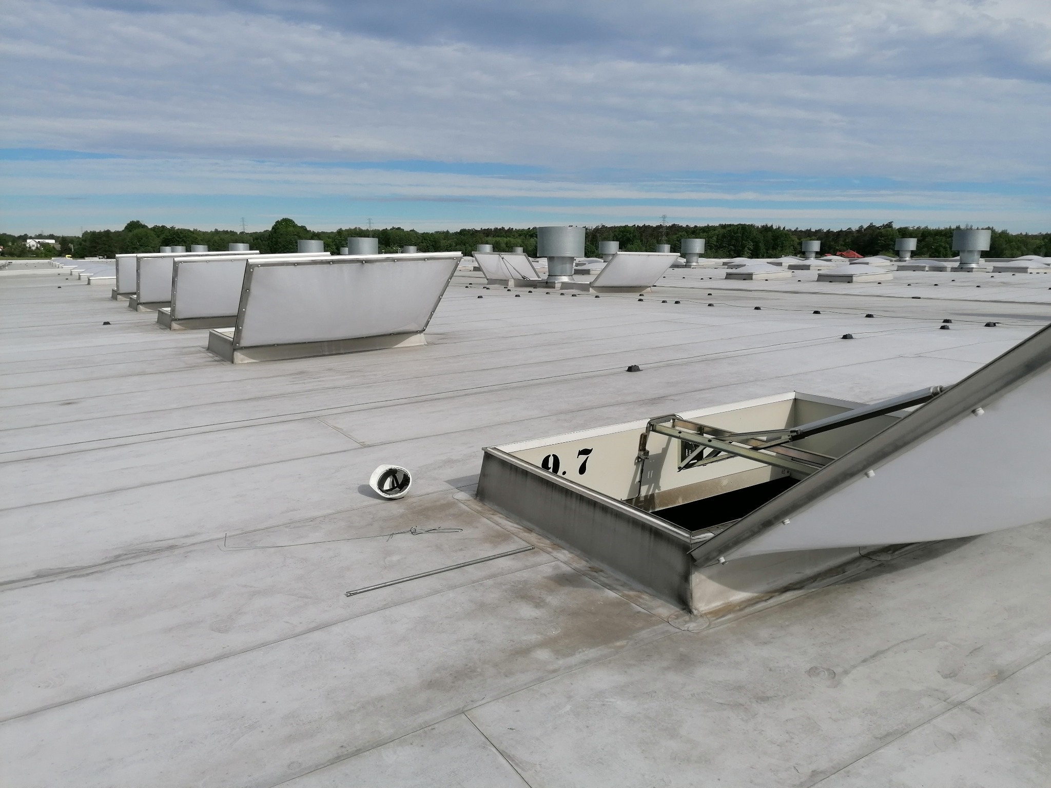 Płaski dach przemysłowy z otwartym świetlikiem dachowym i kaskiem ochronnym obok, w tle widoczne inne świetliki i kominy wentylacyjne.
