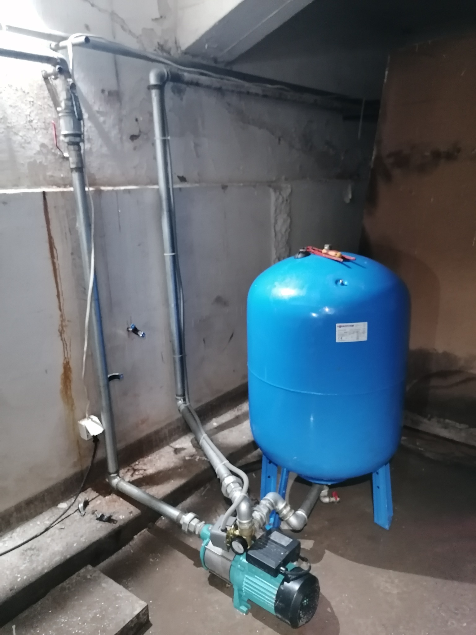 Niebieski zbiornik hydroforowy z podłączoną pompą i rurami w piwnicy, na zbiorniku leżą czerwone szczypce.