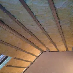 Poddasze z widoczną izolacją z wełny mineralnej między metalowymi profilami, częściowo zakryta folią aluminiową, z oknem dachowym po lewej stronie.