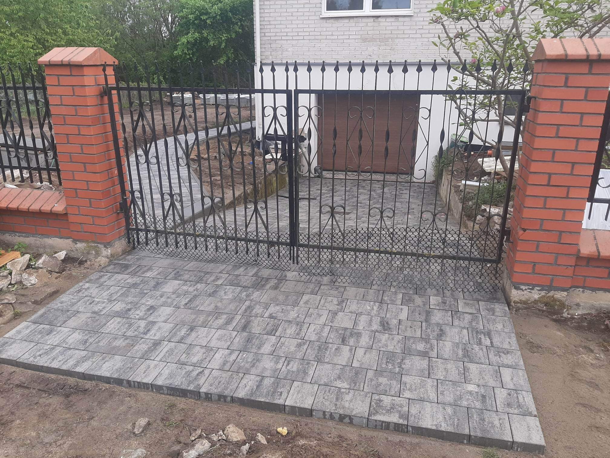 Ułożona szara kostka brukowa prowadząca do metalowej bramy z ceglanymi słupkami, widok na podjazd i garaż w tle.