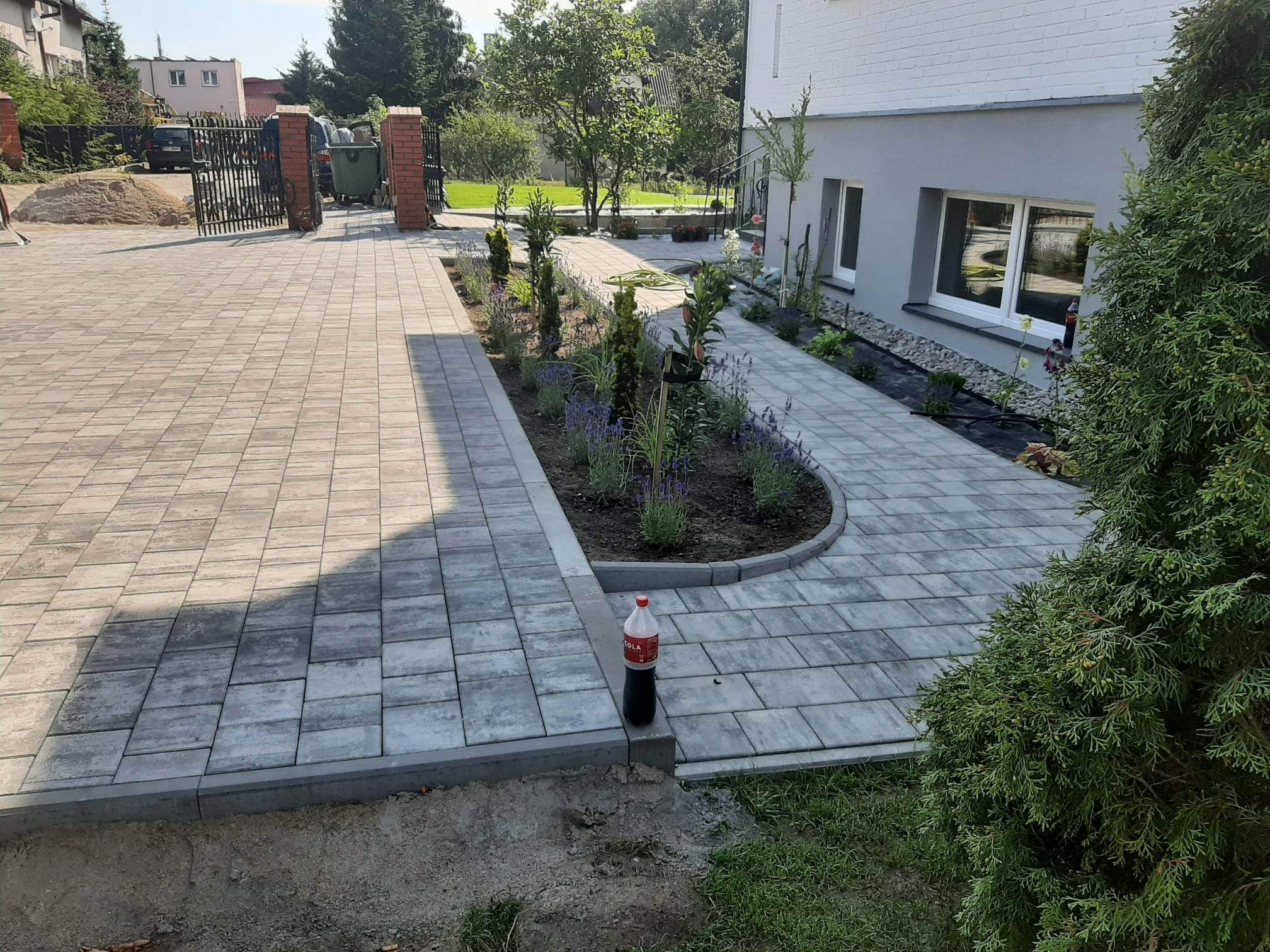 Świeżo ułożona, szara kostka brukowa tworząca ścieżkę i podjazd w Sulęczynie, z rabatami kwiatowymi po bokach i butelką coli stojącą na krawężniku.