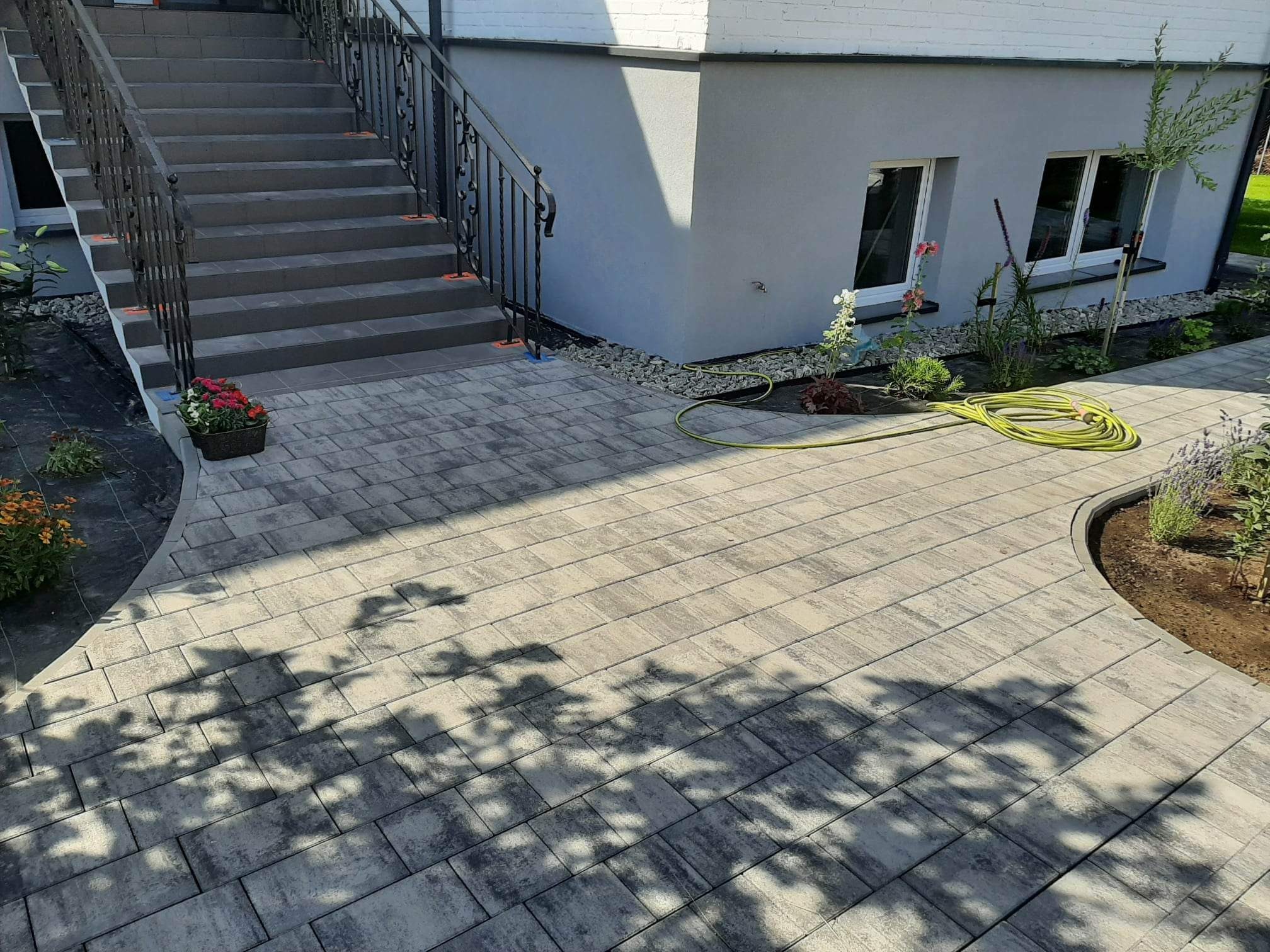 Ułożona kostka brukowa w odcieniach szarości przed budynkiem z betonowymi schodami i metalową balustradą, widoczny wąż ogrodowy i rabaty z roślinami.