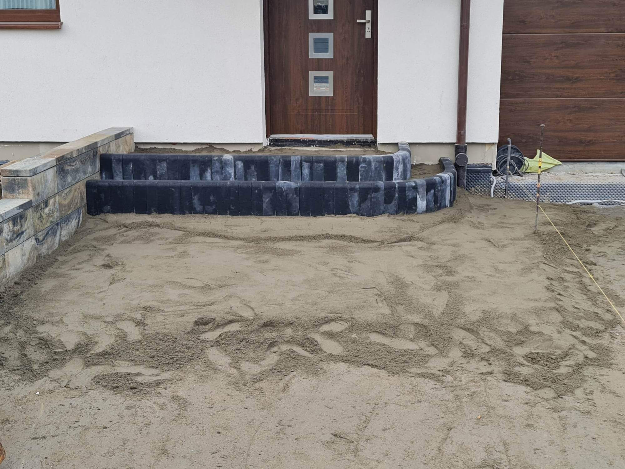 Stopnie wejściowe do domu w trakcie budowy z czarnej kostki obrzeżowej, piasek rozsypany na podłożu, widoczna brązowa brama garażowa i drzwi wejściowe.