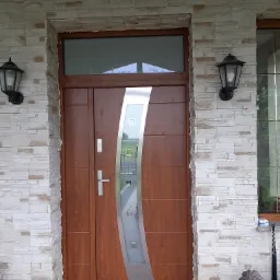 Brązowe drzwi wejściowe z przeszkleniem w srebrnej ramie, zamontowane w elewacji z jasnego kamienia, oświetlone dwoma czarnymi lampami zewnętrznymi.