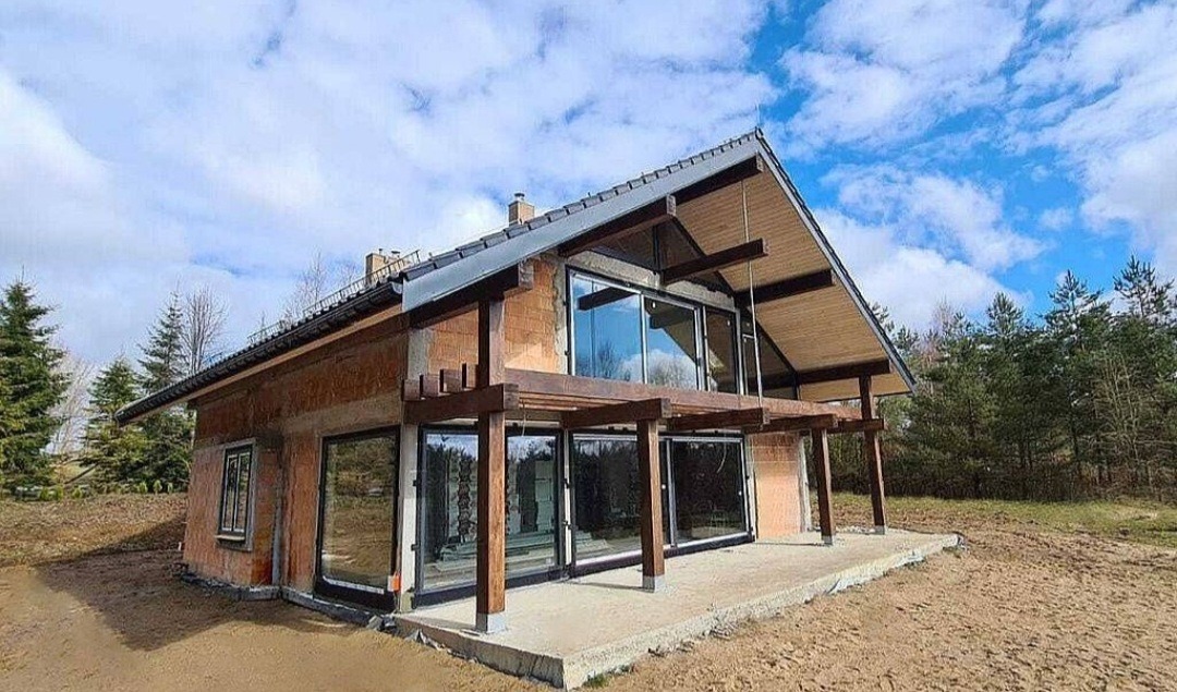 Nowoczesny dom jednorodzinny w trakcie budowy, z dużymi przeszkleniami i drewnianą konstrukcją tarasu, na tle błękitnego nieba z chmurami.
