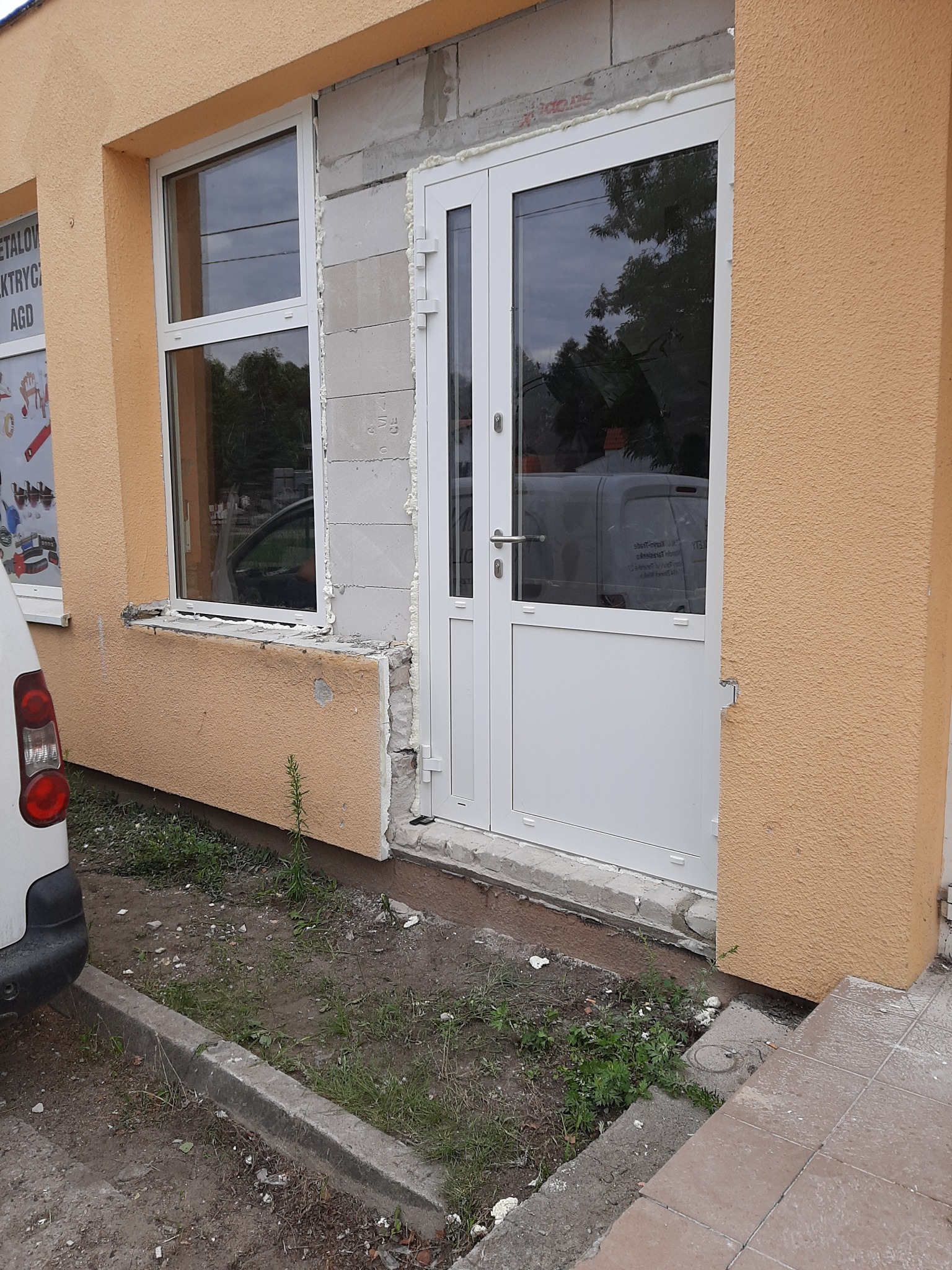 Montaż białych okien i drzwi wejściowych w budynku z otynkowaną elewacją w kolorze piaskowym, widoczne ślady pianki montażowej i niedokończone wykończenie wokół okna i drzwi.
