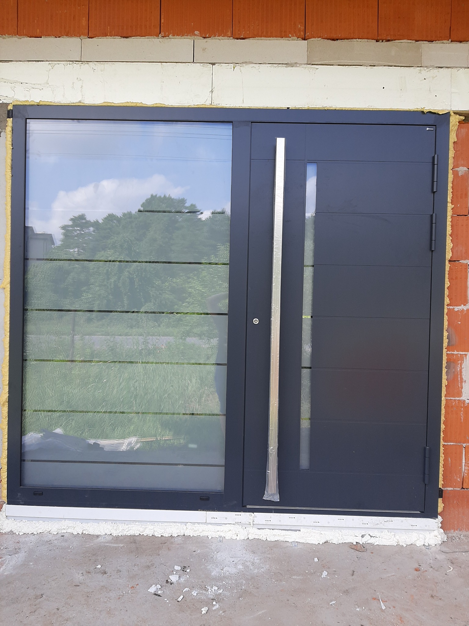 Nowoczesne, grafitowe drzwi wejściowe z bocznym przeszkleniem, zamontowane w surowym stanie budynku, widoczne pianka montażowa i cegła.