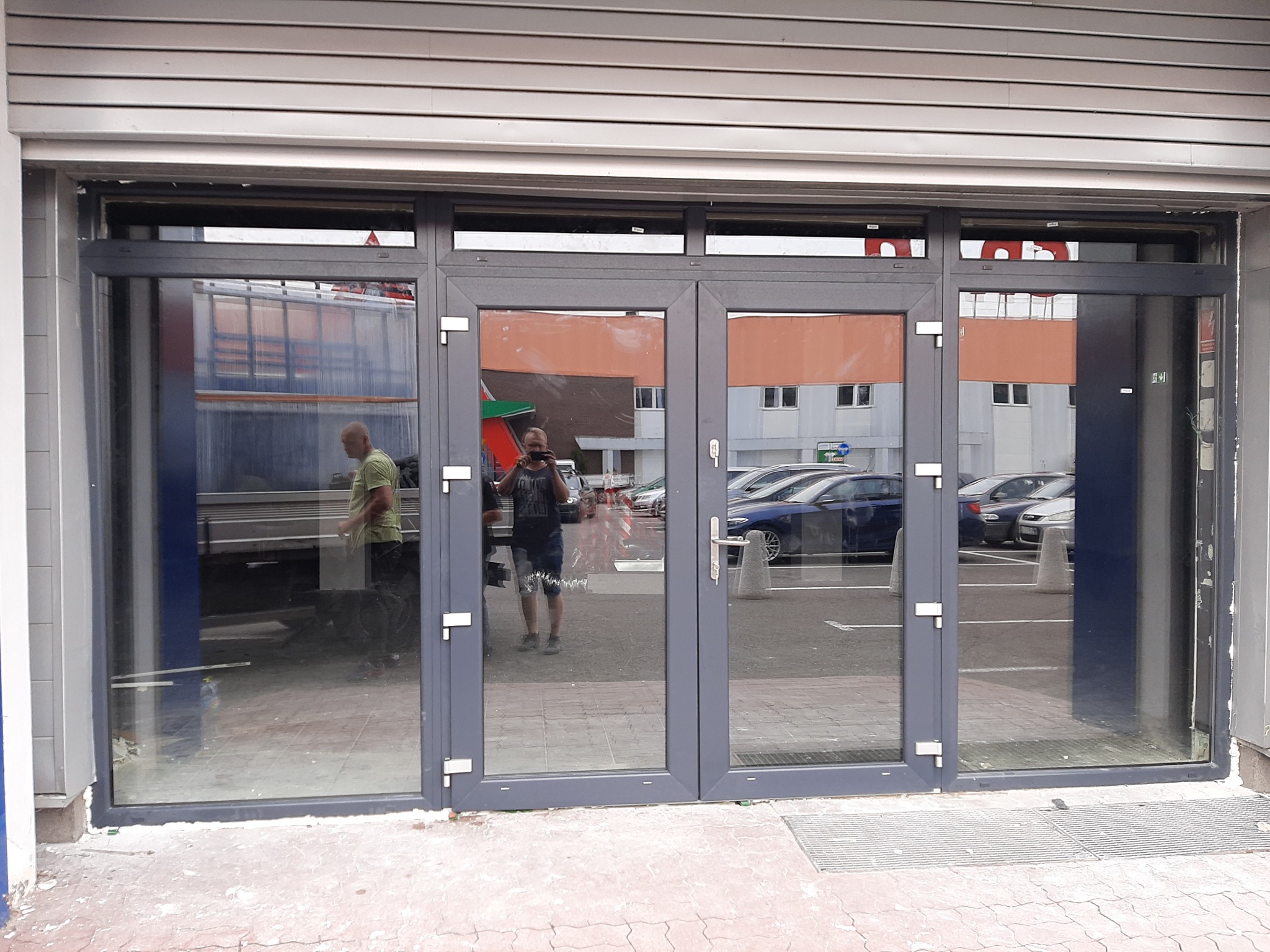 Szare, dwuskrzydłowe drzwi wejściowe z dużymi przeszkleniami, widoczne z zewnątrz, z odbiciem osoby robiącej zdjęcie oraz fragmentu zaparkowanych samochodów i ciężarówki w szybach.