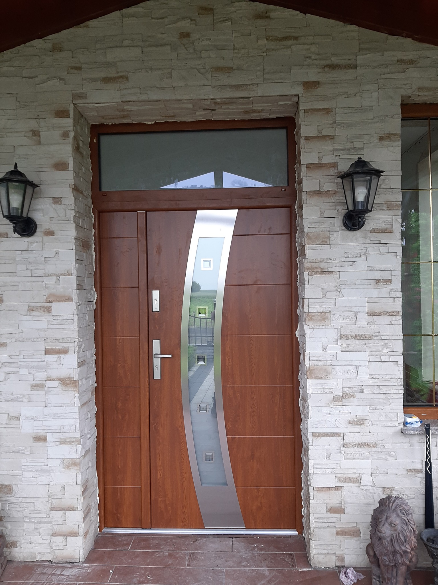 Brązowe drzwi wejściowe z przeszkleniem w srebrnej ramie, zamontowane w elewacji z jasnego kamienia, oświetlone dwoma czarnymi lampami zewnętrznymi.