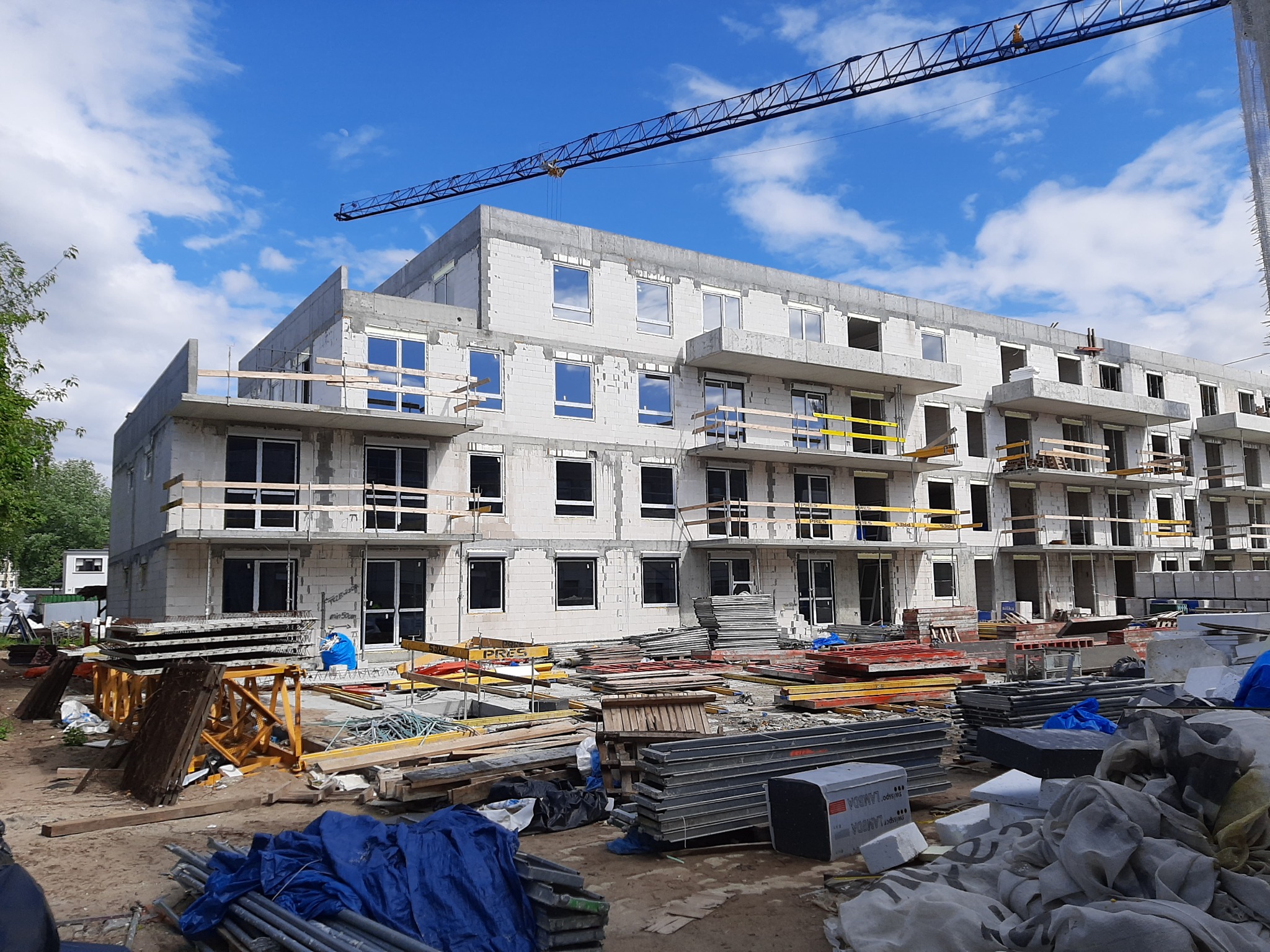 Budynek wielorodzinny w trakcie budowy, widoczne okna z PCV, rusztowania i żuraw budowlany na tle błękitnego nieba z chmurami.
