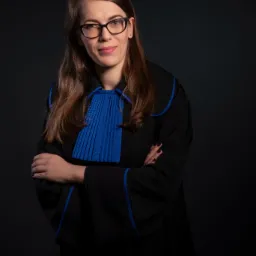 Portret kobiety w todze z niebieskim żabotem i lamówką, z założonymi rękoma na ciemnym tle.