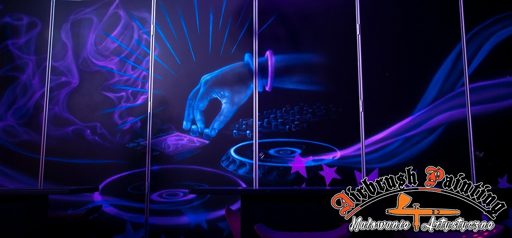 Fluorescencyjna grafika na ścianie przedstawiająca dłoń DJ-a miksującego muzykę na gramofonach, wykonana techniką airbrush, z elementami fioletowych smug i gwiazd.