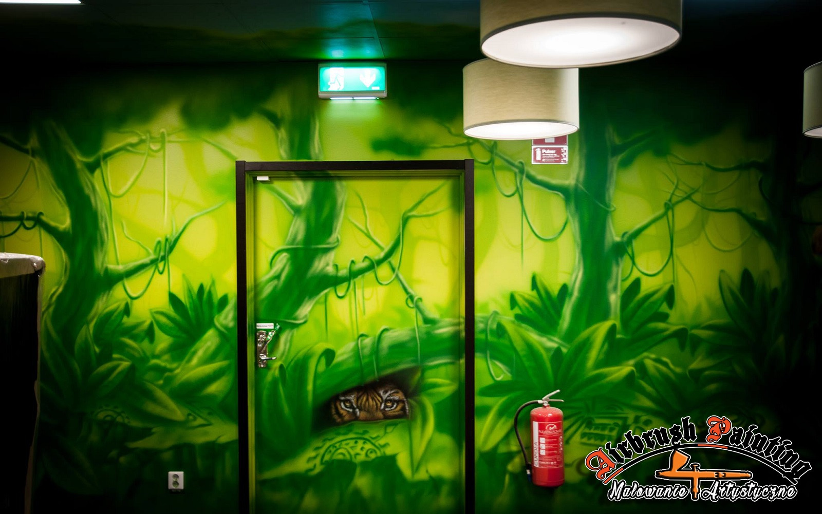 Wnętrze z zielonym malunkiem dżungli na ścianie i drzwiach, z ukrytym w zaroślach wzorem oczu tygrysa na drzwiach, gaśnica w prawym dolnym rogu.