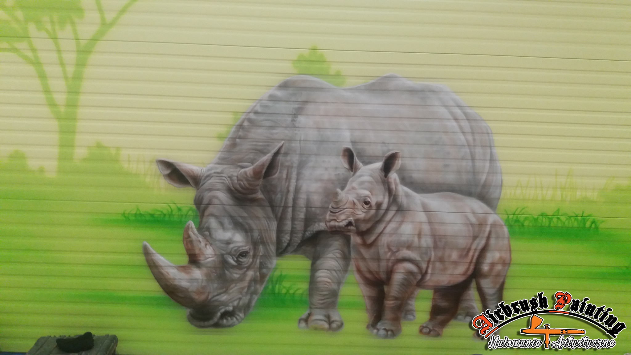 Ilustracja nosorożca z młodym na metalowej ścianie garażu, wykonana techniką airbrush, w tle zarys drzewa.
