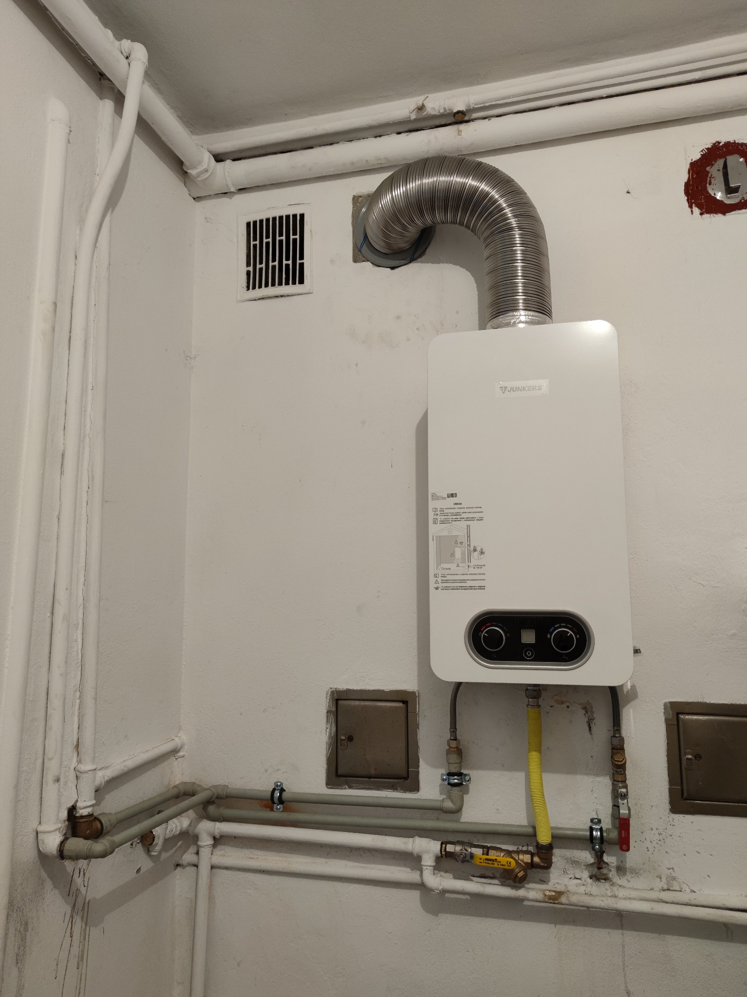 Biały piec gazowy Junkers z podłączonymi rurami, w tym żółtą rurą gazową, zamontowany na pomalowanej na biało ścianie w rogu pomieszczenia z widocznymi innymi rurami i kratką wentylacyjną.