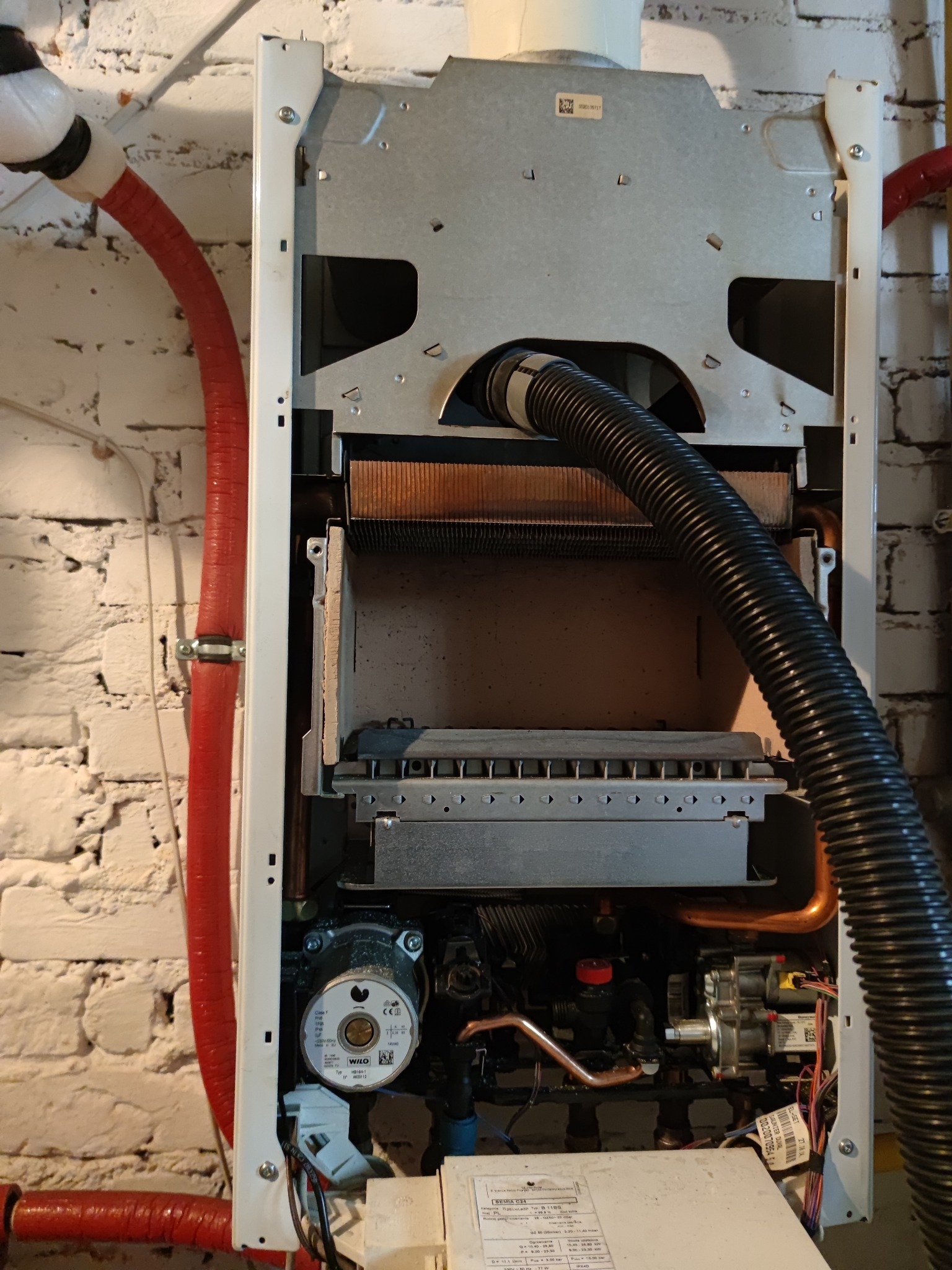 Otwarty piec gazowy z widocznymi elementami wewnętrznymi, rurami miedzianymi i czarnym karbowanym wężem, zamontowany na ścianie z cegły. Widoczna pompa obiegowa WILO.