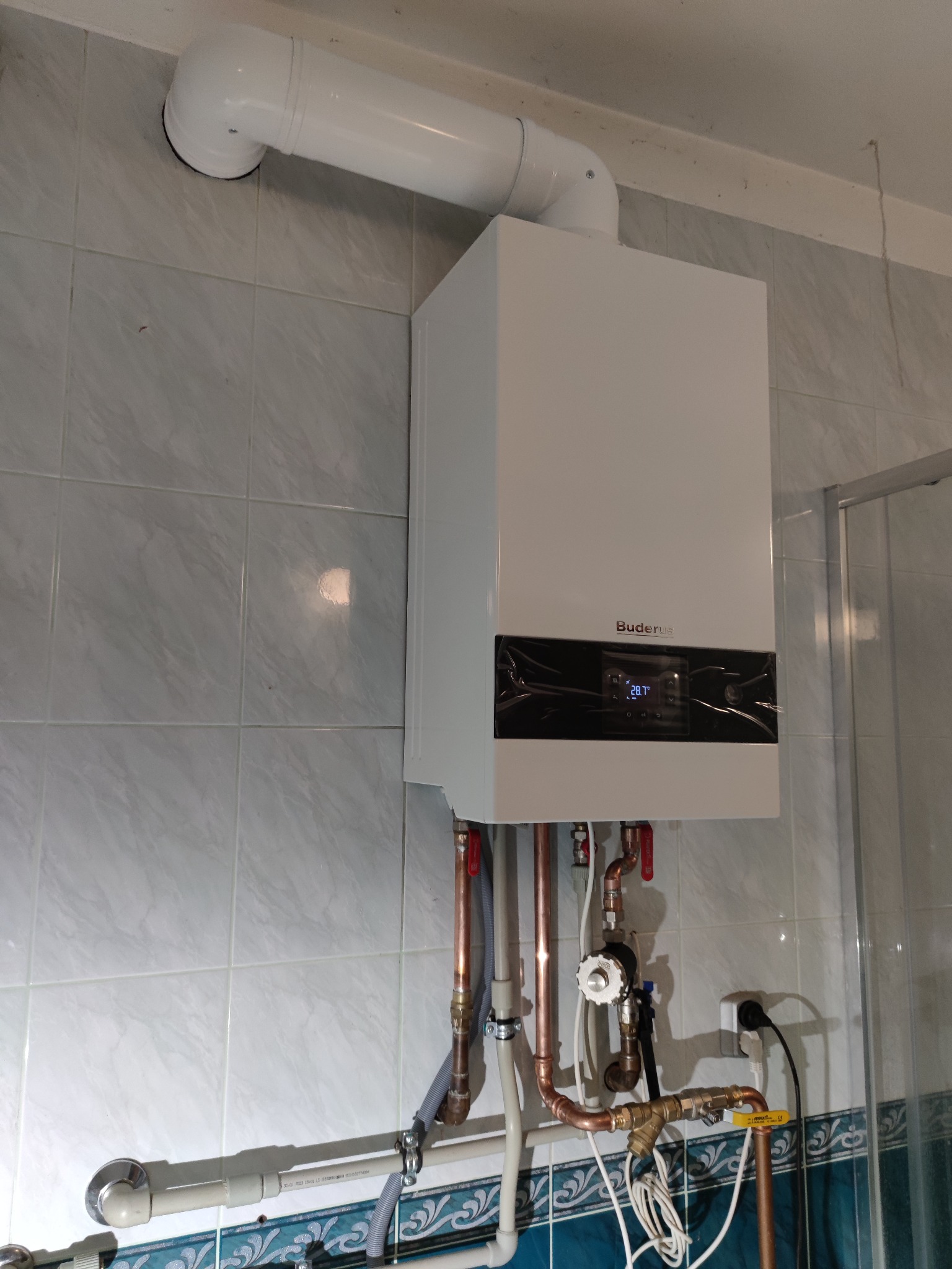 Biały, wiszący piec gazowy marki Buderus z podłączonymi miedzianymi rurami i białym kominem na tle jasnych, kwadratowych płytek ceramicznych, widoczny fragment kabiny prysznicowej.
