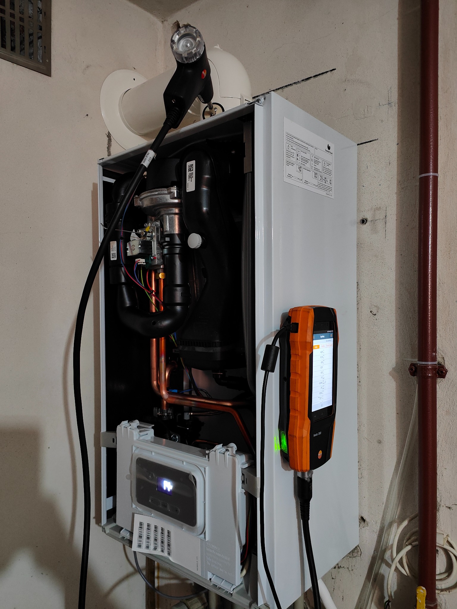 Biały piec gazowy wiszący na ścianie, podłączony do analizatora spalin Testo 300; widoczne miedziane rury i elementy wewnętrzne kotła.