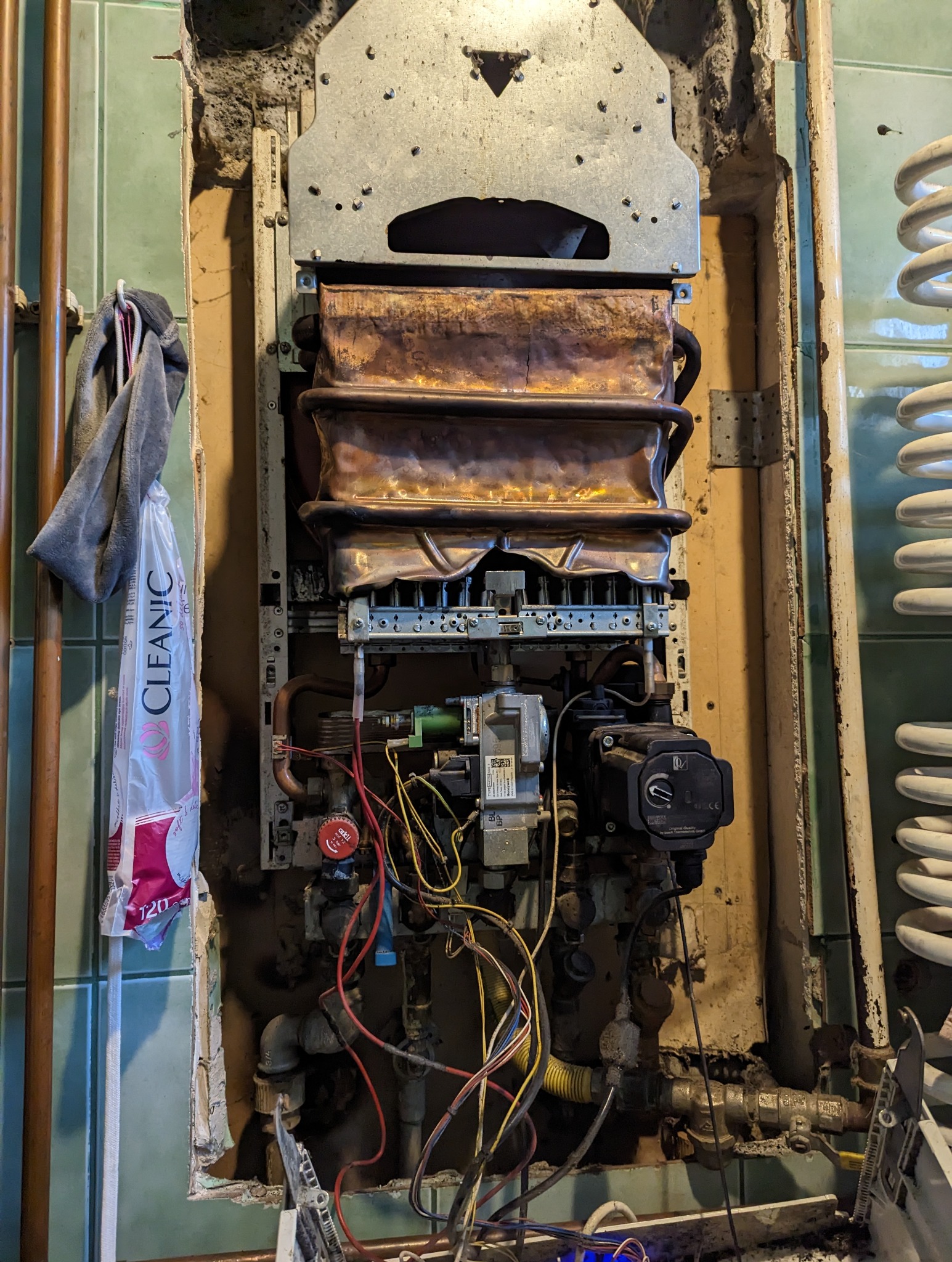 Odkryty piec gazowy z widocznymi elementami wewnętrznymi, przewodami i rurami, zamontowany na ścianie z zielonymi płytkami, obok torba reklamowa i grzejnik spiralny.