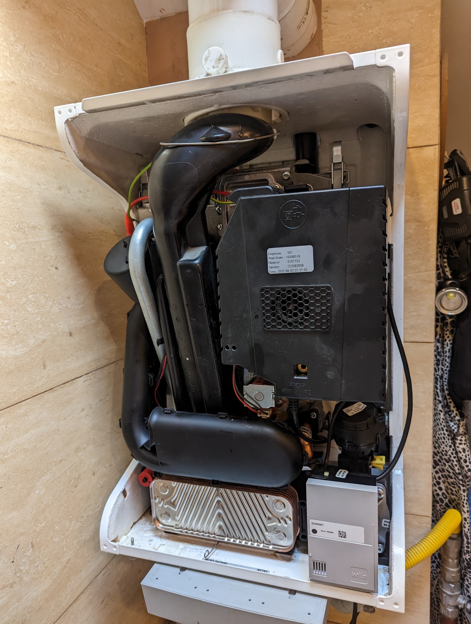 Wnętrze otwartego, białego pieca gazowego z widocznymi elementami instalacji, w tym czarnymi rurami, srebrnym wymiennikiem ciepła i żółtym przewodem gazowym, na tle beżowych płytek.