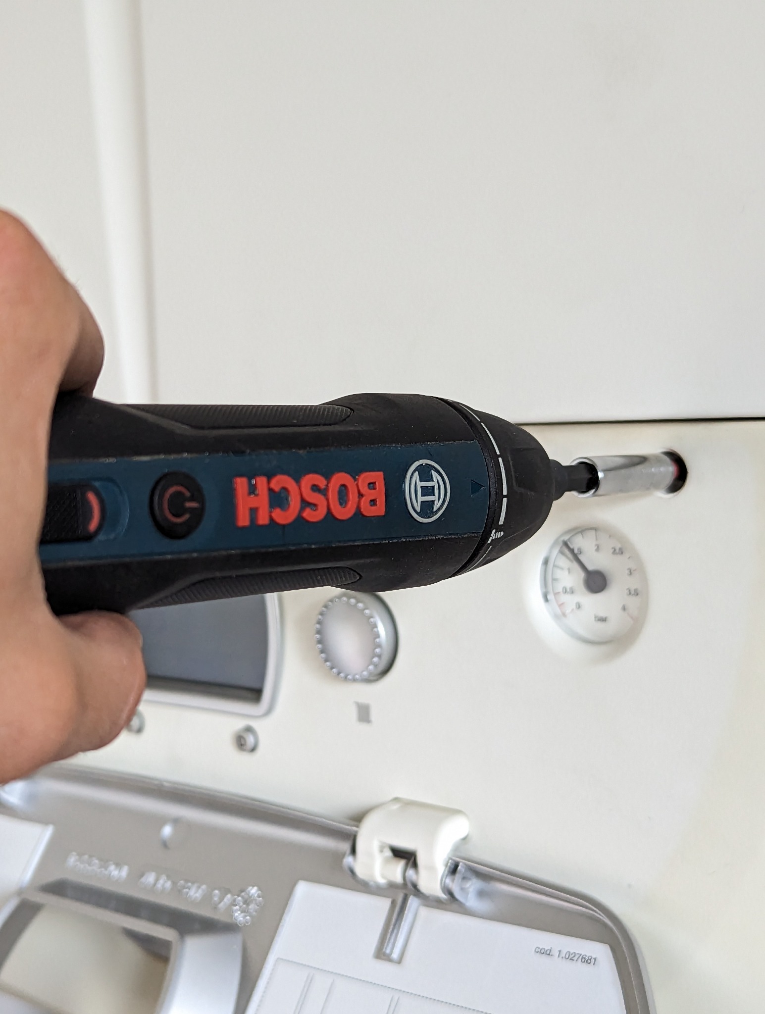 Czarny wkrętak akumulatorowy Bosch w dłoni wkręca śrubę w obudowę pieca gazowego z widocznym manometrem.