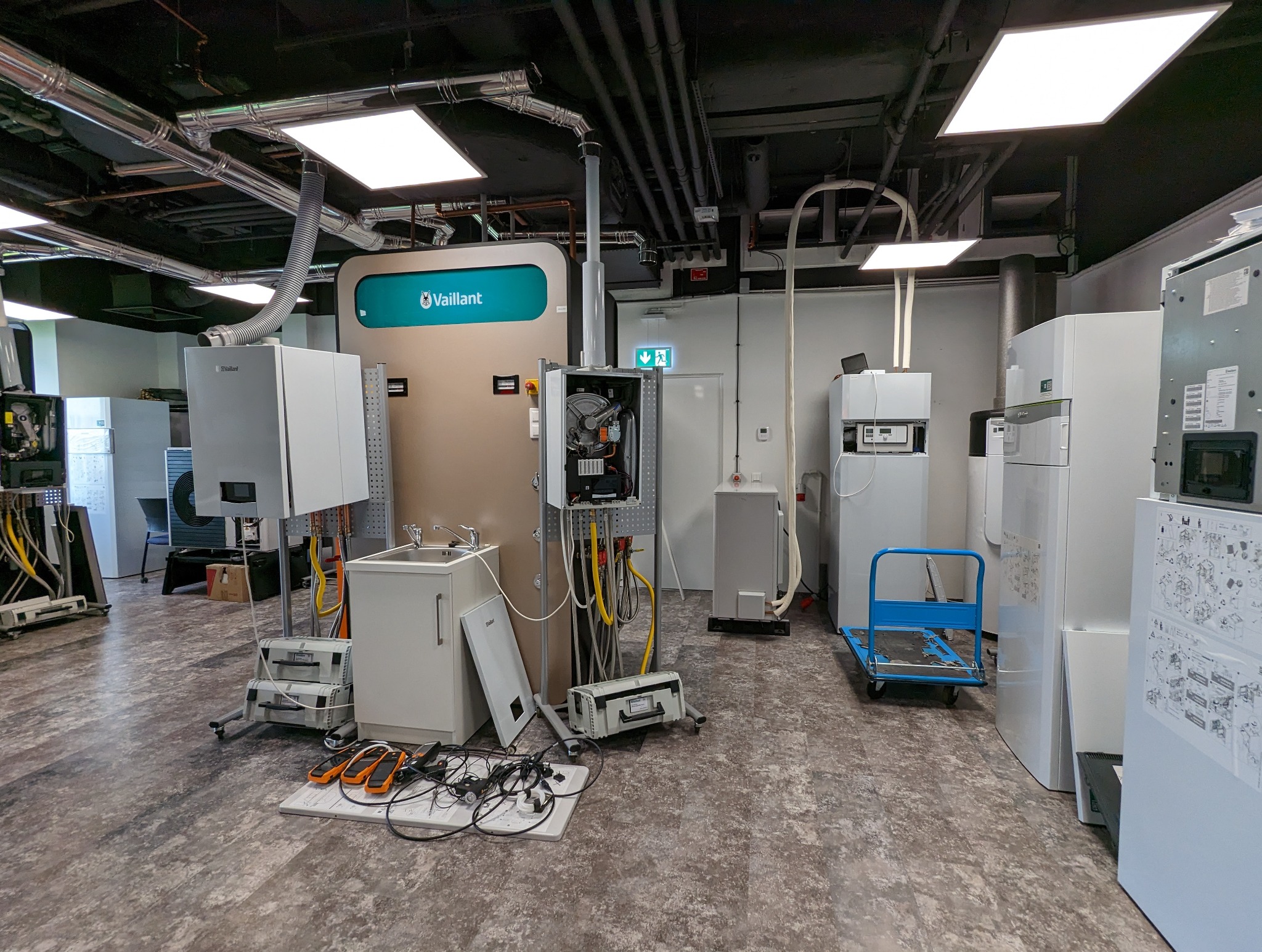 Sala szkoleniowa z piecami gazowymi marki Vaillant, narzędziami i stanowiskami do nauki montażu i serwisu, widoczne otwarte obudowy pieców, instalacje i schematy.