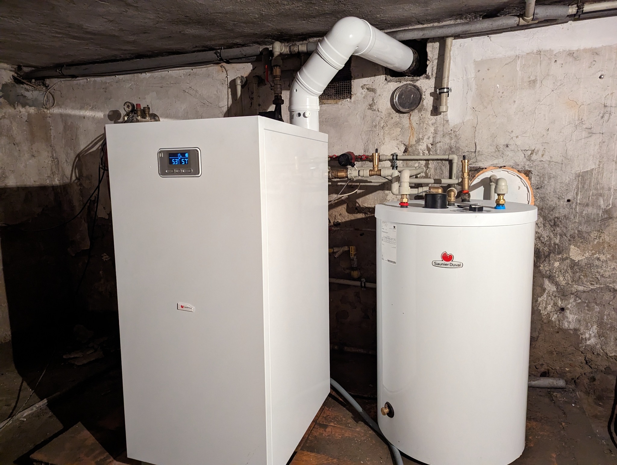 Nowoczesny, biały piec gazowy kondensacyjny z podgrzewaczem wody marki Saunier Duval, zainstalowany w piwnicy o surowych, betonowych ścianach. Widoczne rury instalacyjne, wyświetlacz pieca pokazuje...