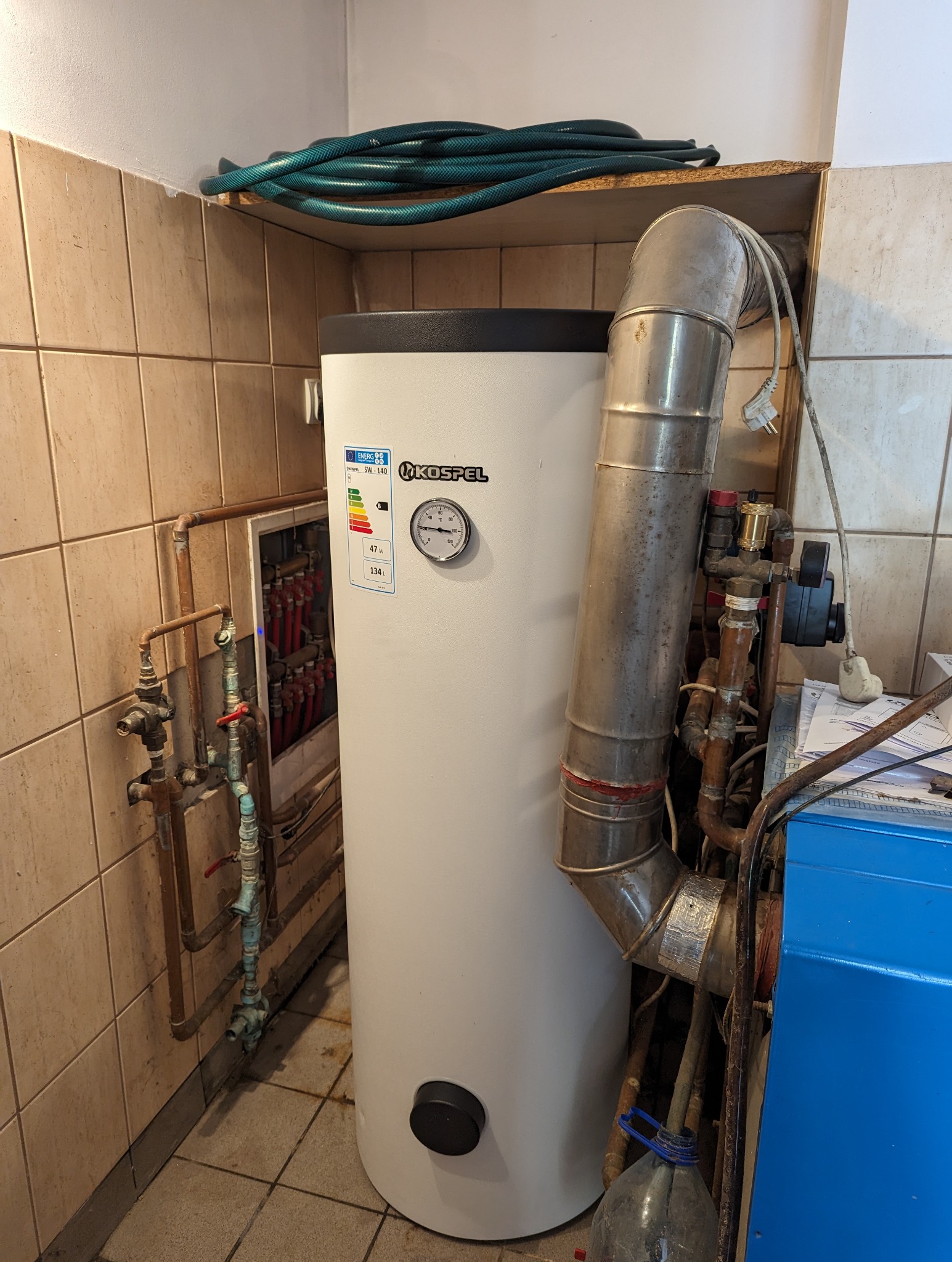 Biały, cylindryczny zasobnik ciepłej wody użytkowej marki Kospel z widocznym wskaźnikiem temperatury i etykietą energetyczną, podłączony do instalacji CO z rurami miedzianymi i stalowymi...