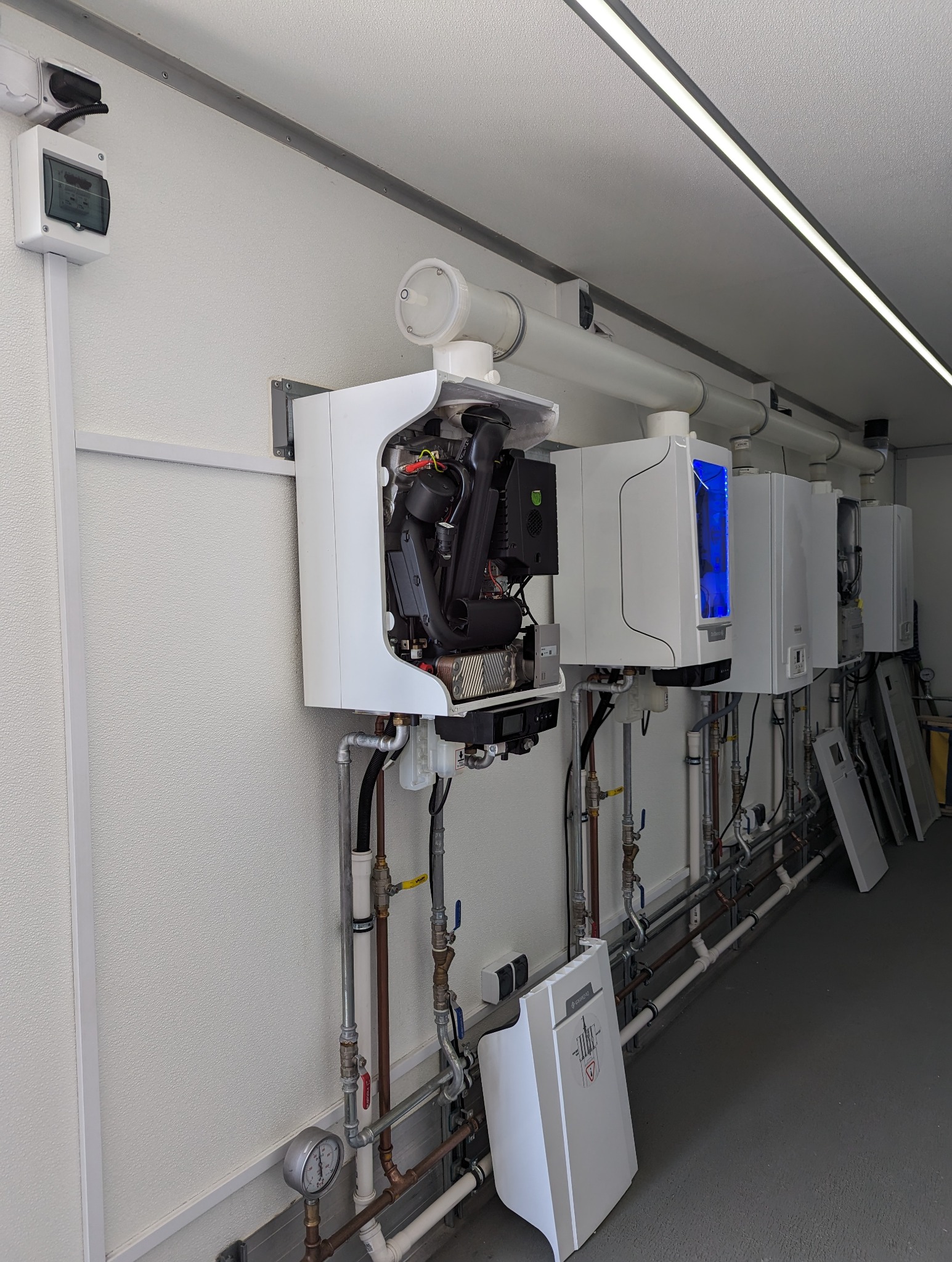 Rząd białych pieców gazowych zamontowanych na ścianie, jeden z otwartą obudową ukazującą wewnętrzne komponenty, rury i zawory pod spodem, w tle widoczne drzwi i oświetlenie sufitowe.