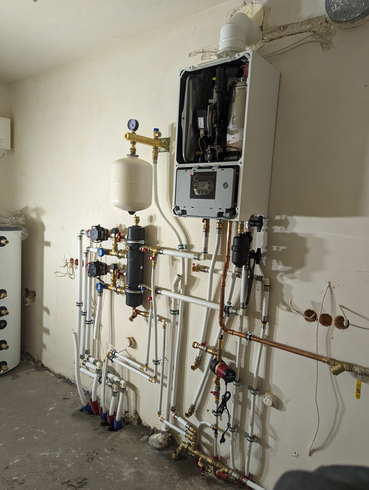 Nowo zainstalowany, wiszący na ścianie kocioł gazowy Buderus z otwartą obudową, widoczne podłączenia hydrauliczne z rur miedzianych i tworzyw sztucznych, zbiornik wyrównawczy oraz grupa pompowa.