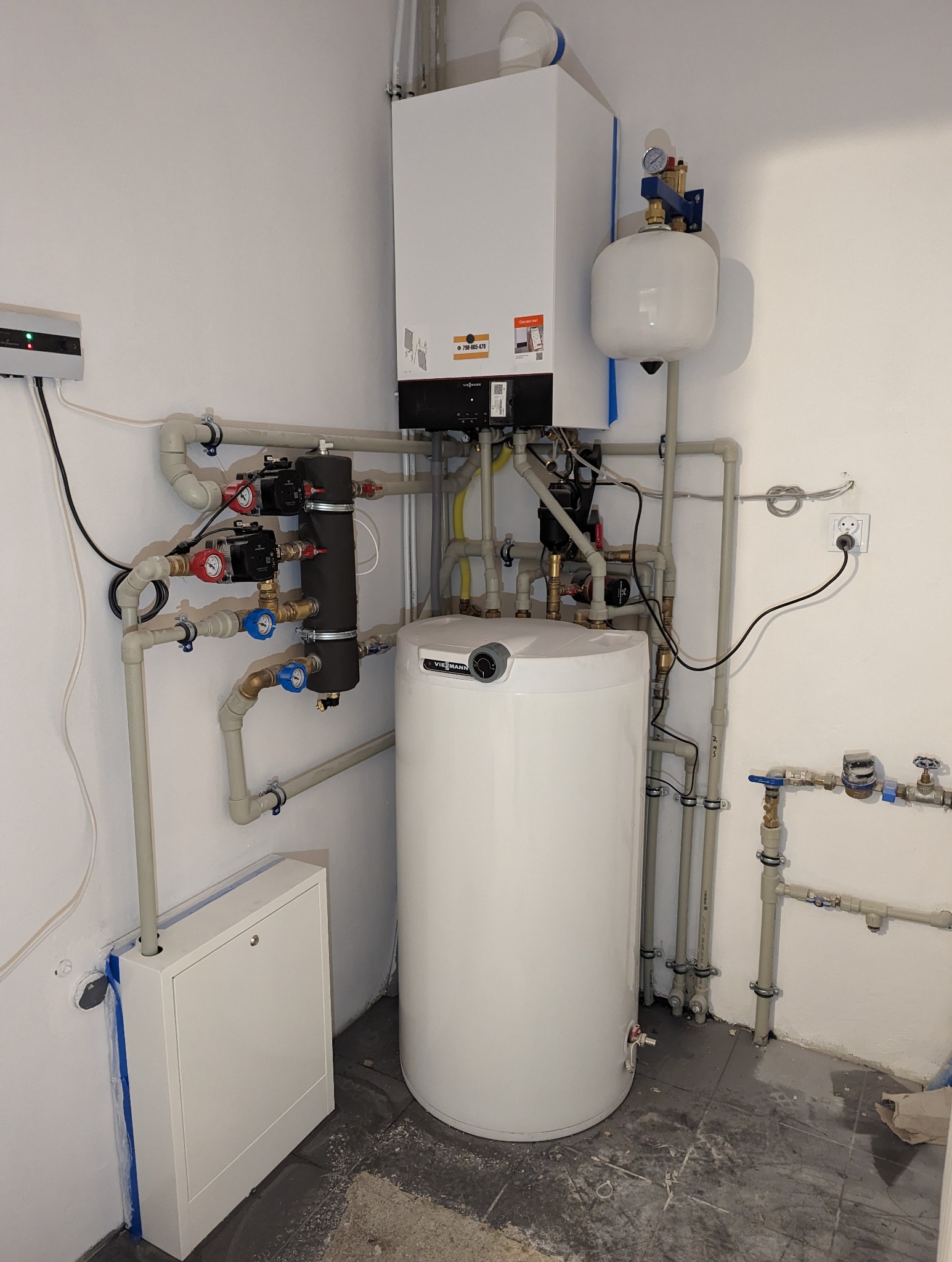 Kompletna instalacja kotła gazowego z zasobnikiem ciepłej wody użytkowej, widoczne liczne rury, zawory i pompy obiegowe, zamontowane na ścianie w pomieszczeniu technicznym.