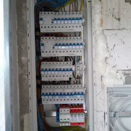 Rozdzielnica elektryczna w trakcie montażu, widoczne kable, bezpieczniki i zabezpieczenia przepięciowe, osadzona w ścianie z surowego betonu z widoczną pianką montażową.