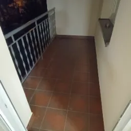 Wąski balkon z brązowymi płytkami podłogowymi, białą balustradą i widokiem na nocne miasto zza balustrady, widziany z wnętrza mieszkania przez otwarte drzwi balkonowe.