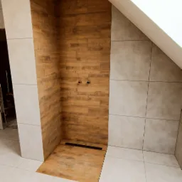 Niedokończona kabina prysznicowa z drewnopodobnymi płytkami, widoczne przyłącza wodne i odpływ liniowy, jasne płytki na podłodze i ścianach, okno dachowe.