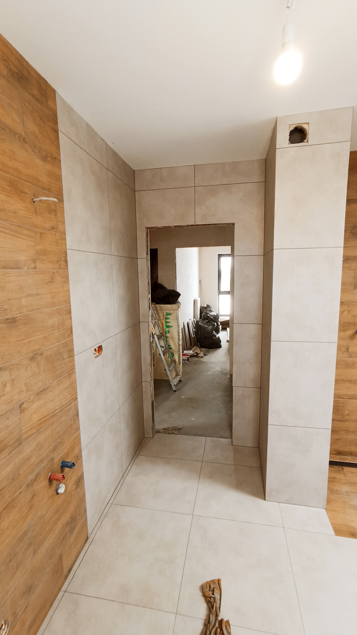 Wnętrze łazienki w trakcie remontu: ściany częściowo wyłożone płytkami w odcieniach beżu i drewna, widoczne przygotowania do montażu armatury, drabina i worki z materiałami budowlanymi w tle.
