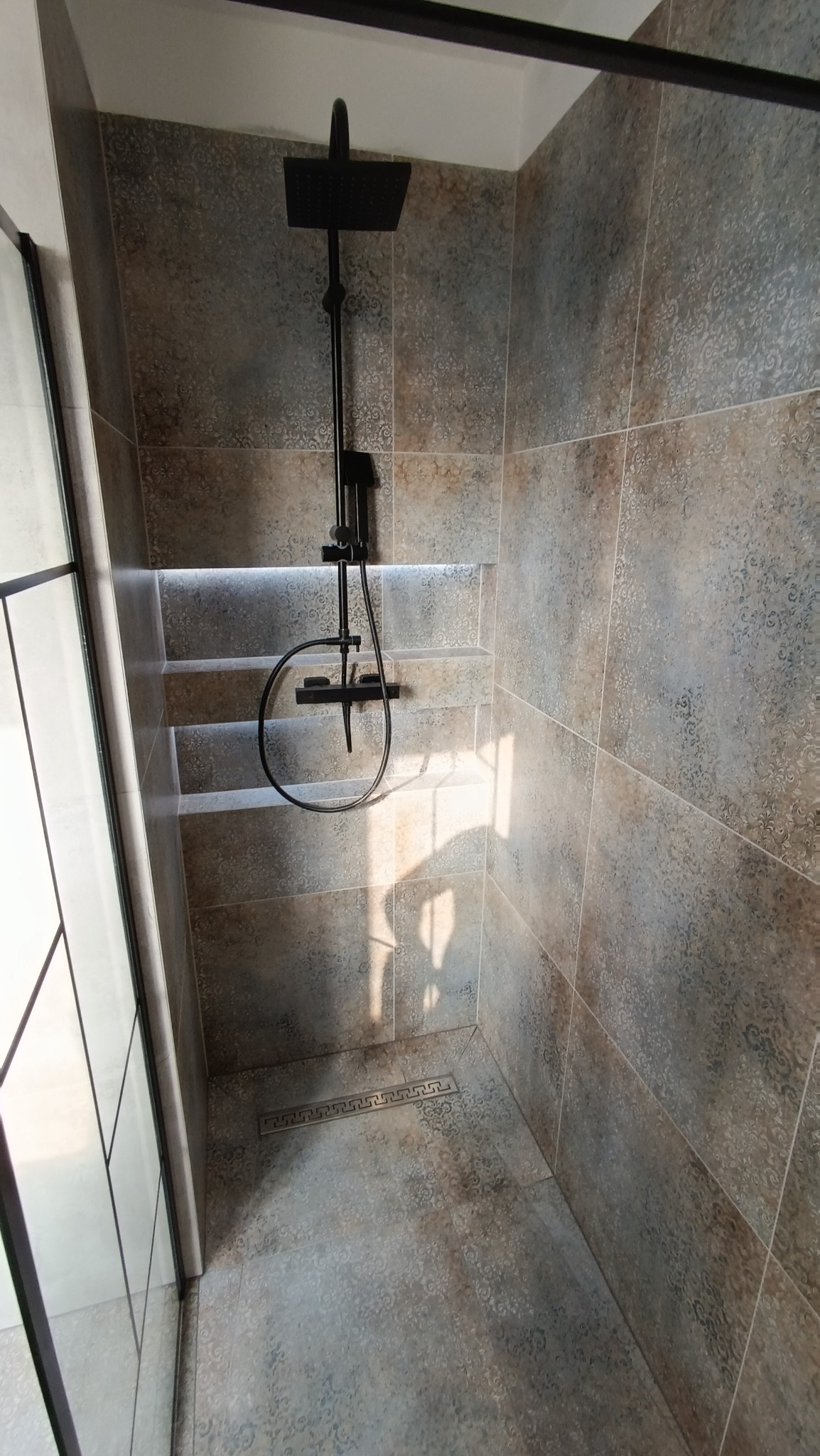 Wykończona kabina prysznicowa z czarną armaturą, płytkami w odcieniach szarości i beżu z delikatnym wzorem oraz wnęką z podświetleniem LED.