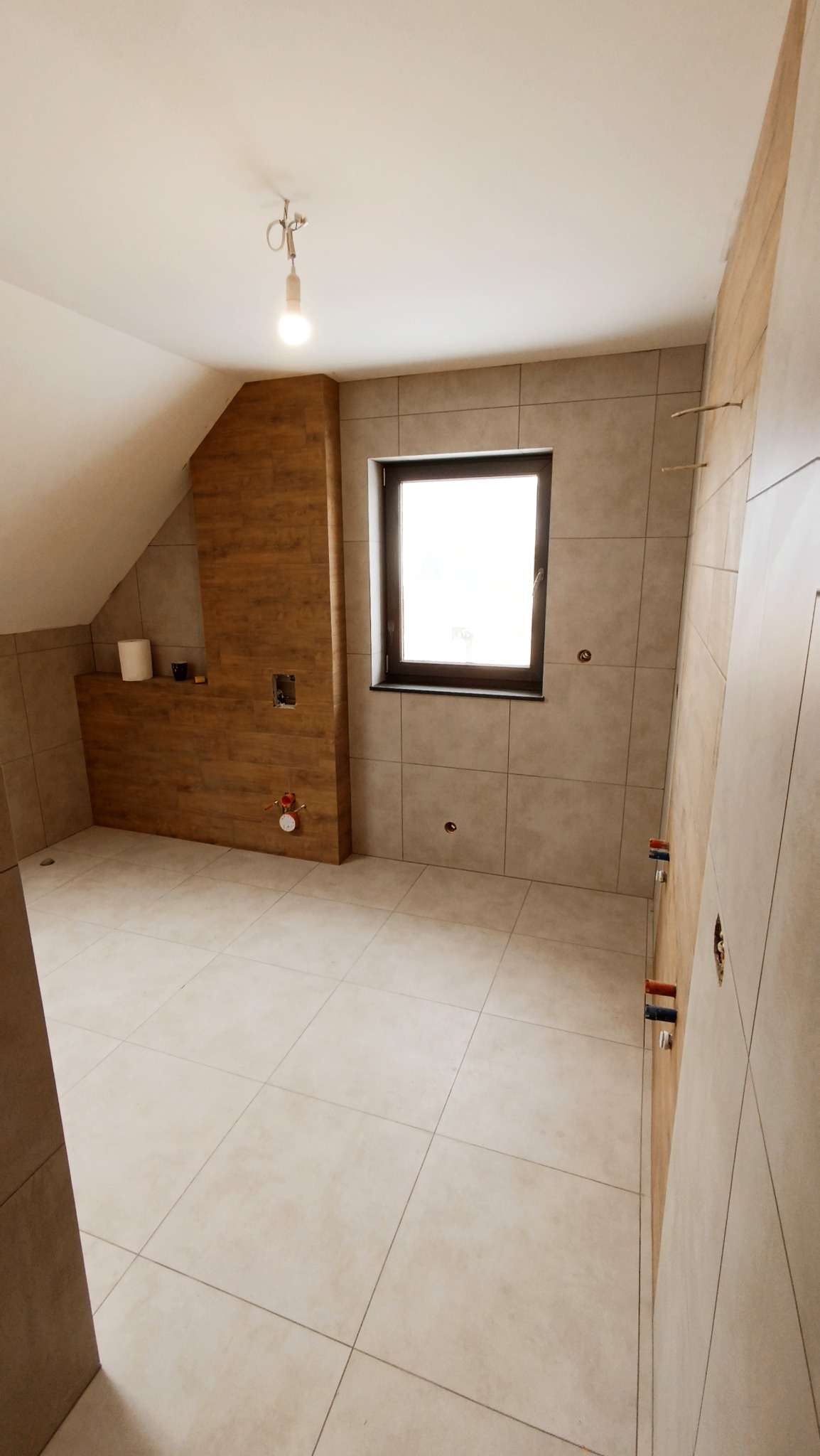 Poddasze łazienki w trakcie wykończenia: beżowe płytki na podłodze i ścianach, drewnopodobna zabudowa, przygotowane punkty instalacyjne, surowe oświetlenie.