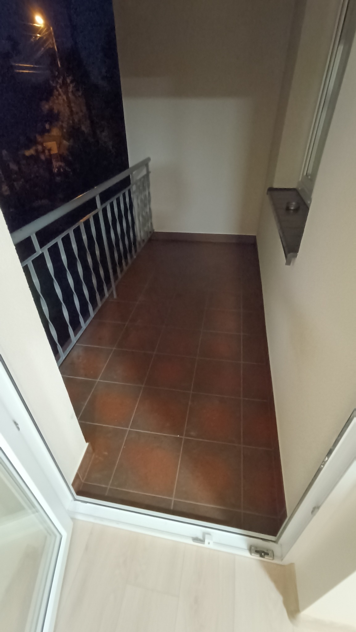 Wąski balkon z brązowymi płytkami podłogowymi, białą balustradą i widokiem na nocne miasto zza balustrady, widziany z wnętrza mieszkania przez otwarte drzwi balkonowe.