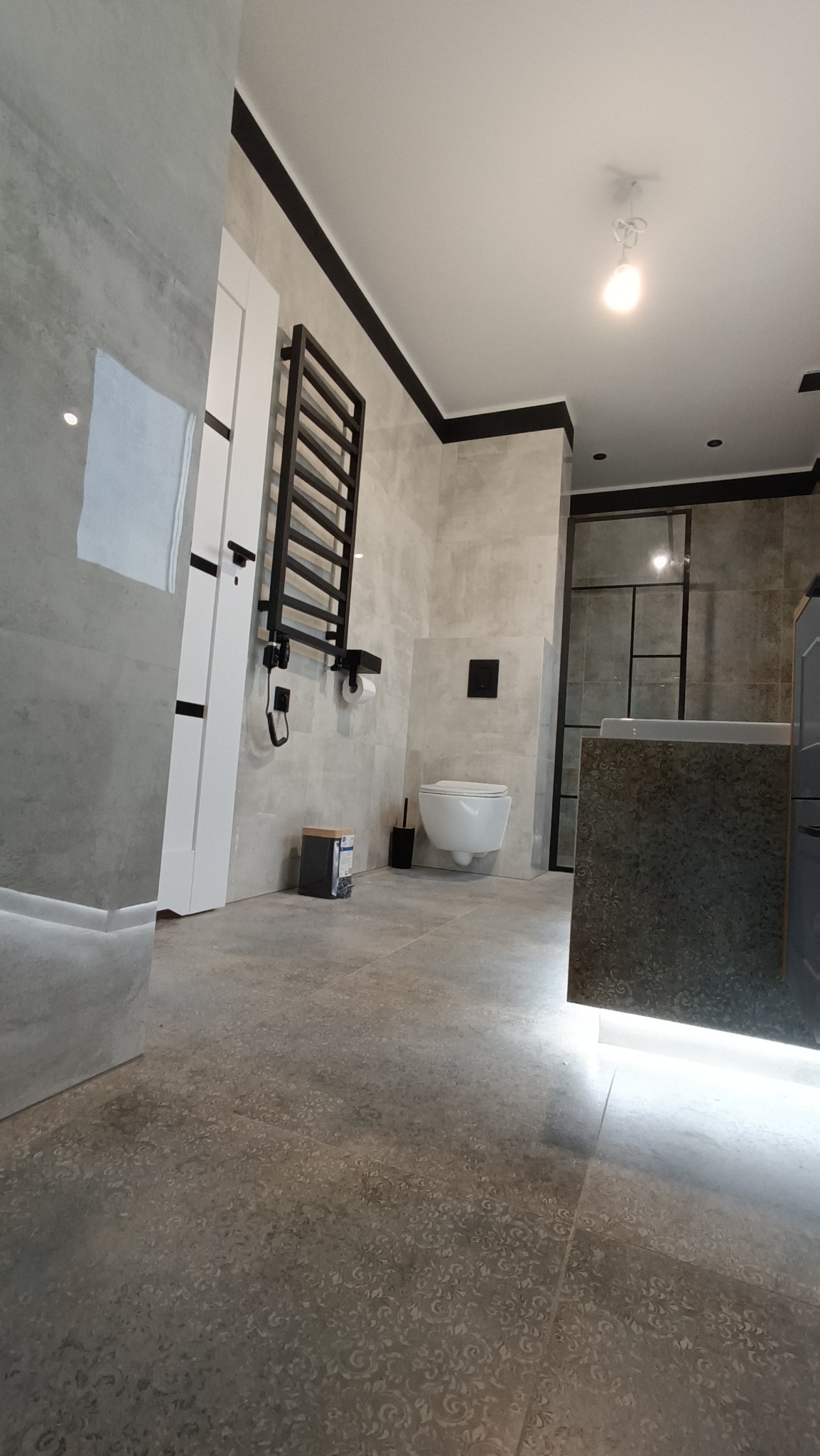 Nowoczesna łazienka wykończona w szarościach, z podwieszaną toaletą, grzejnikiem drabinkowym w kolorze czarnym i kabiną prysznicową z czarnymi ramami. Na podłodze płytki z delikatnym wzorem.