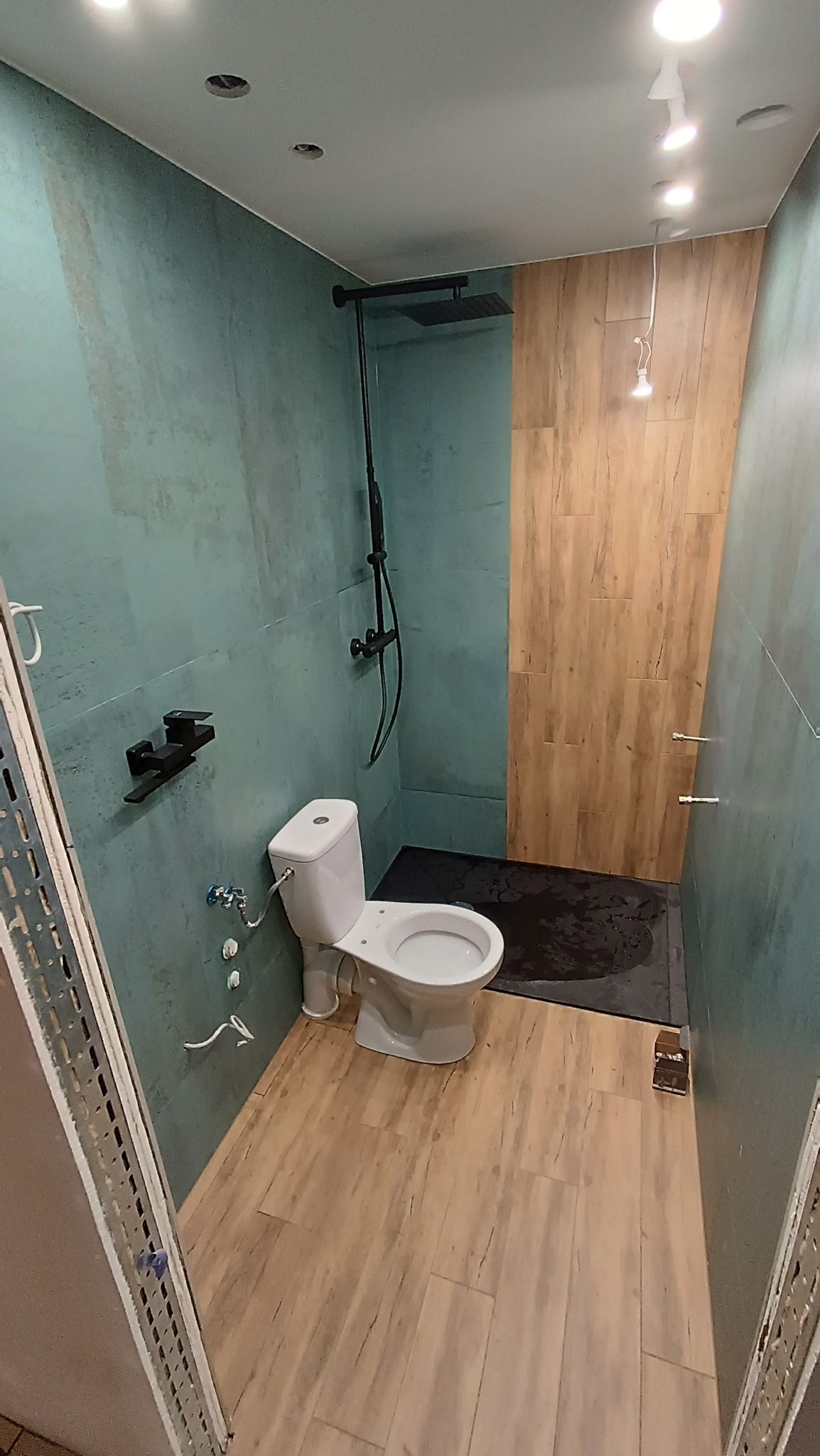 Nowoczesna łazienka z turkusowymi ścianami, drewnopodobną podłogą i czarną armaturą prysznicową typu deszczownica, widoczny biały sedes i fragment odpływu liniowego.